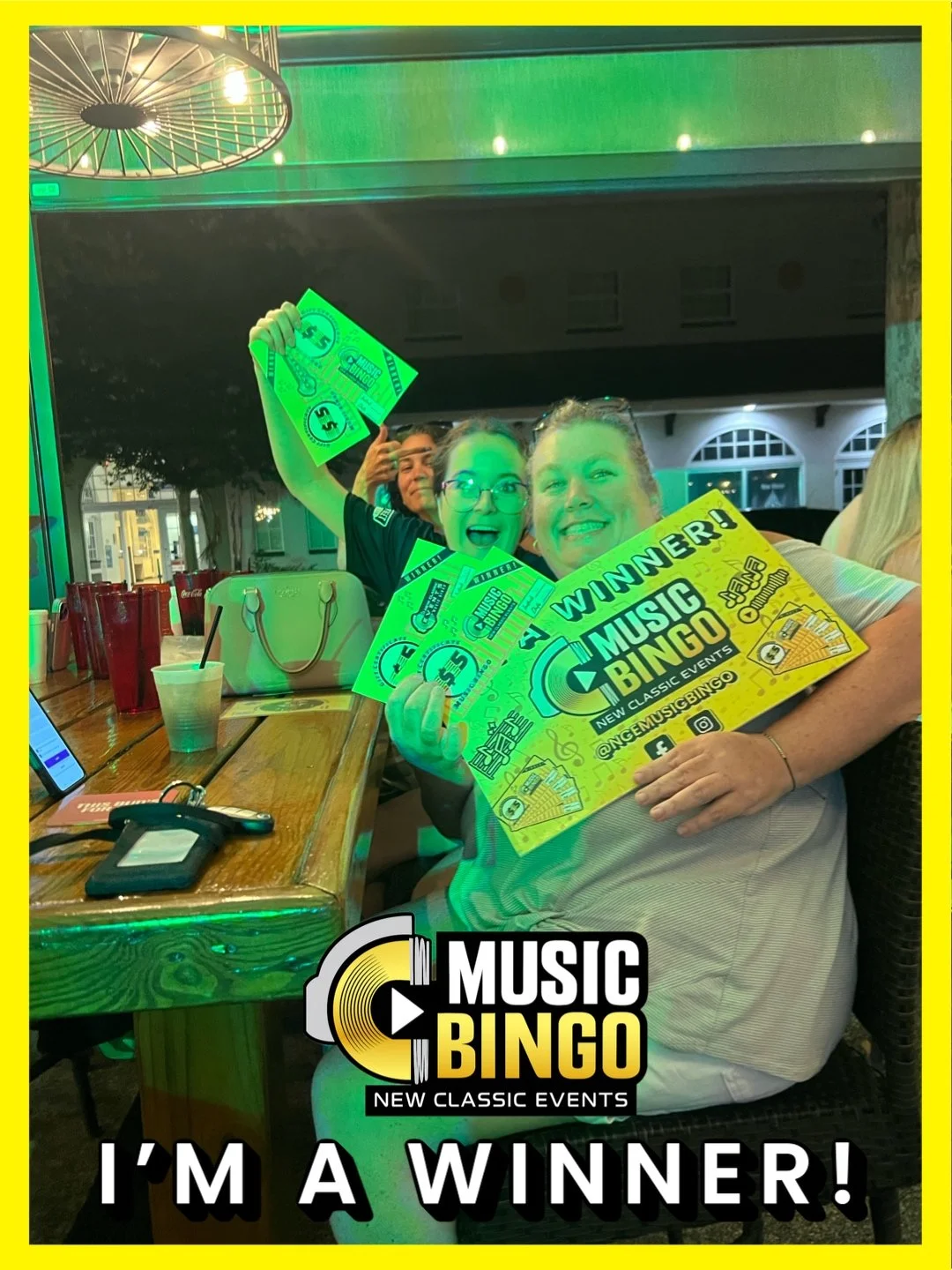 Two women are celebrating at a Music Bingo event, holding a sign that says 'WINNER!', with one woman also holding several green tickets or certificates. They are smiling and appear excited, seated at a table with drinks and personal items, in a livel