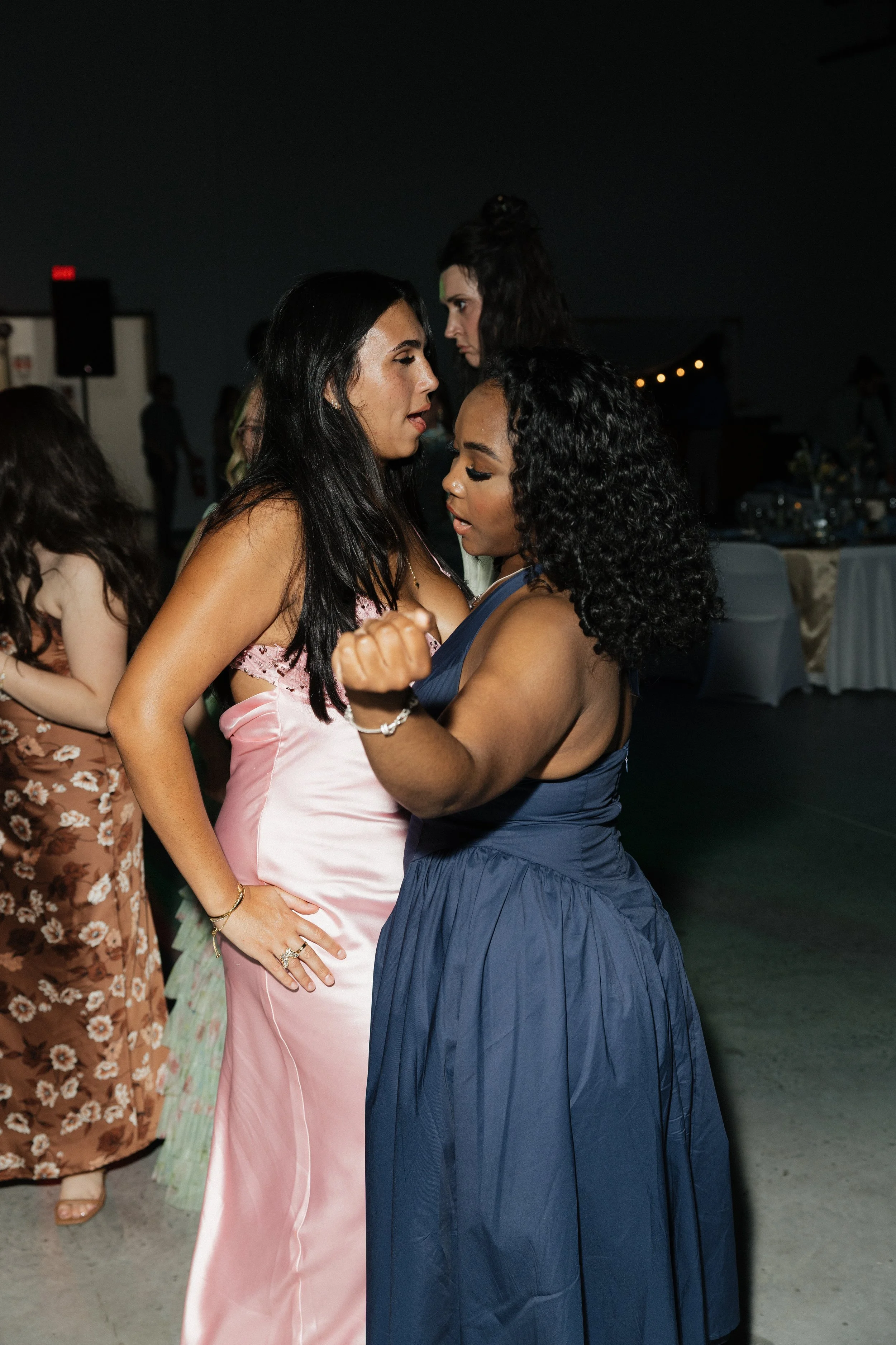 Two women dancing closely at a celebration, with a third woman in the background, on a darkened indoor dance floor.