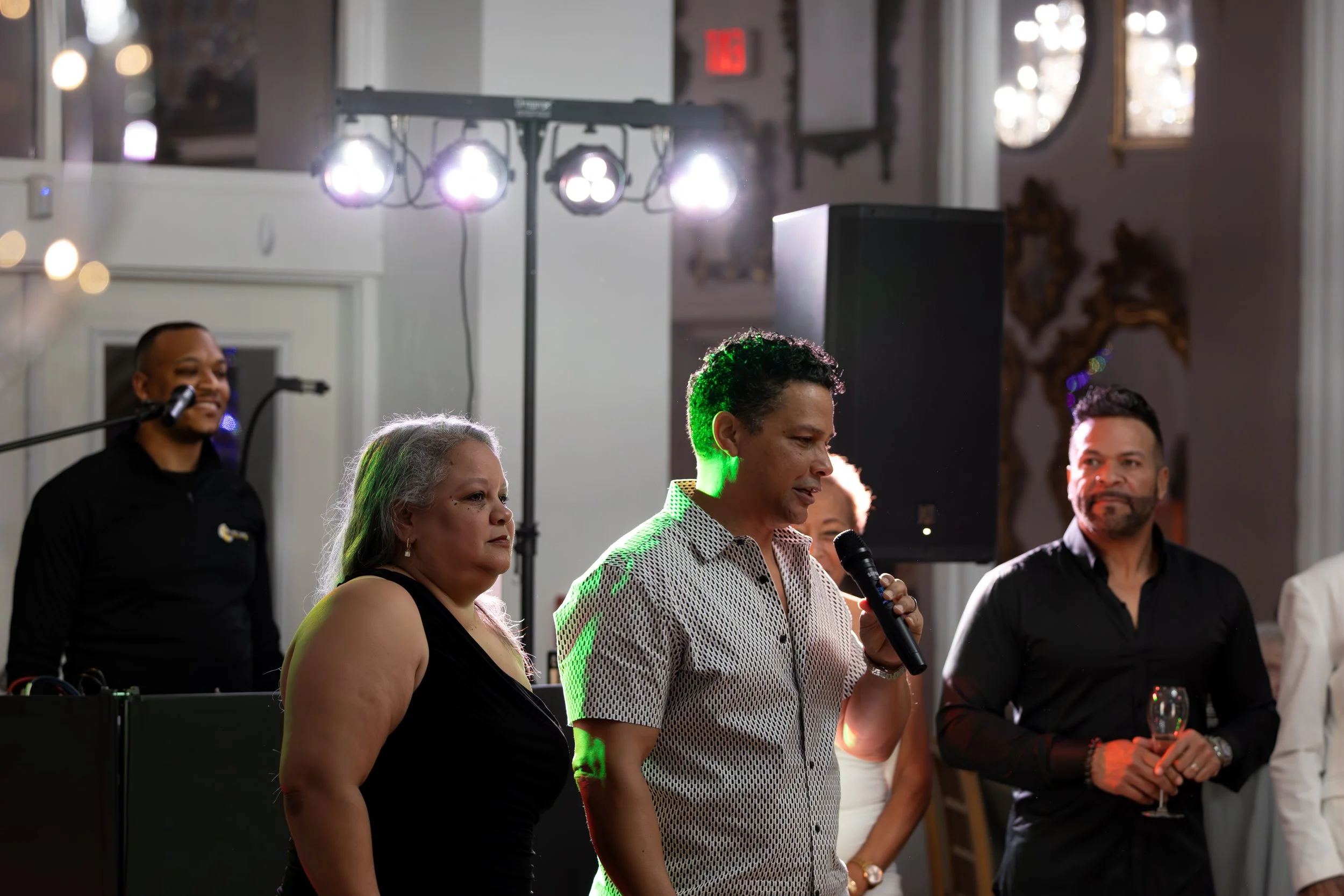 A woman holding a microphone speaking at a formal event, with three other people standing nearby and a DJ in the background.