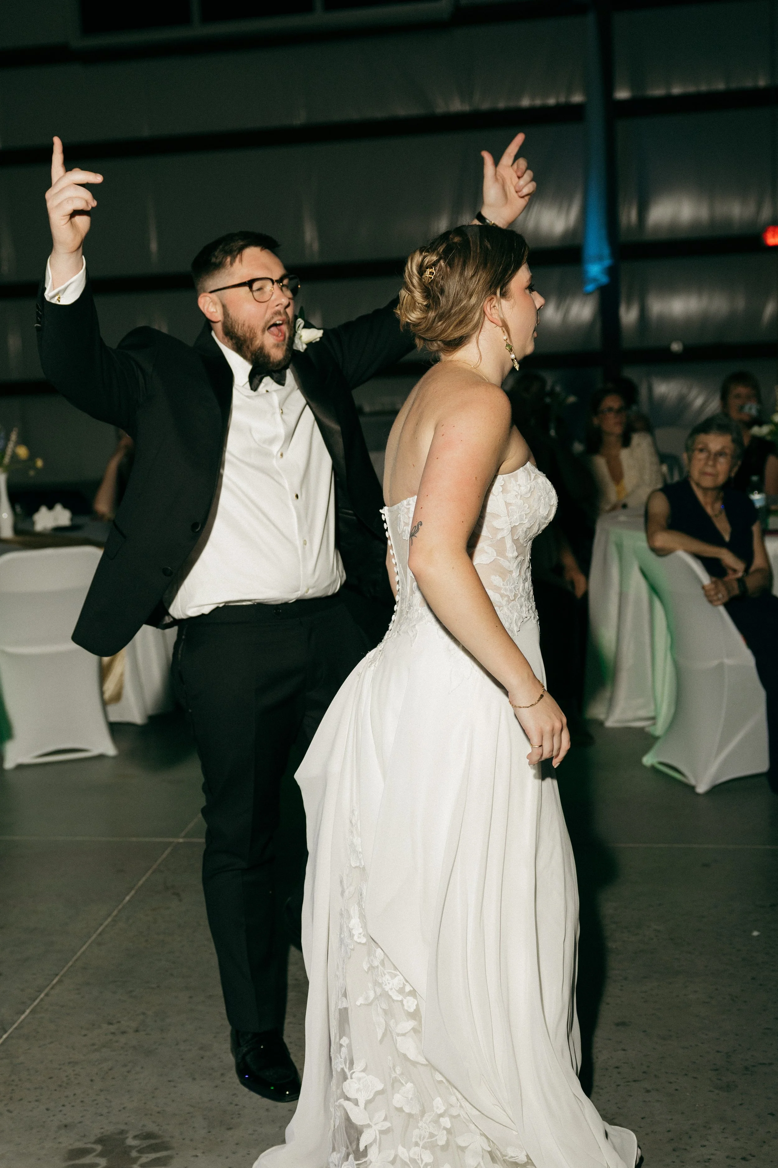 A man and woman dancing at a wedding reception, with the man raising his arms and the woman in a white wedding dress.