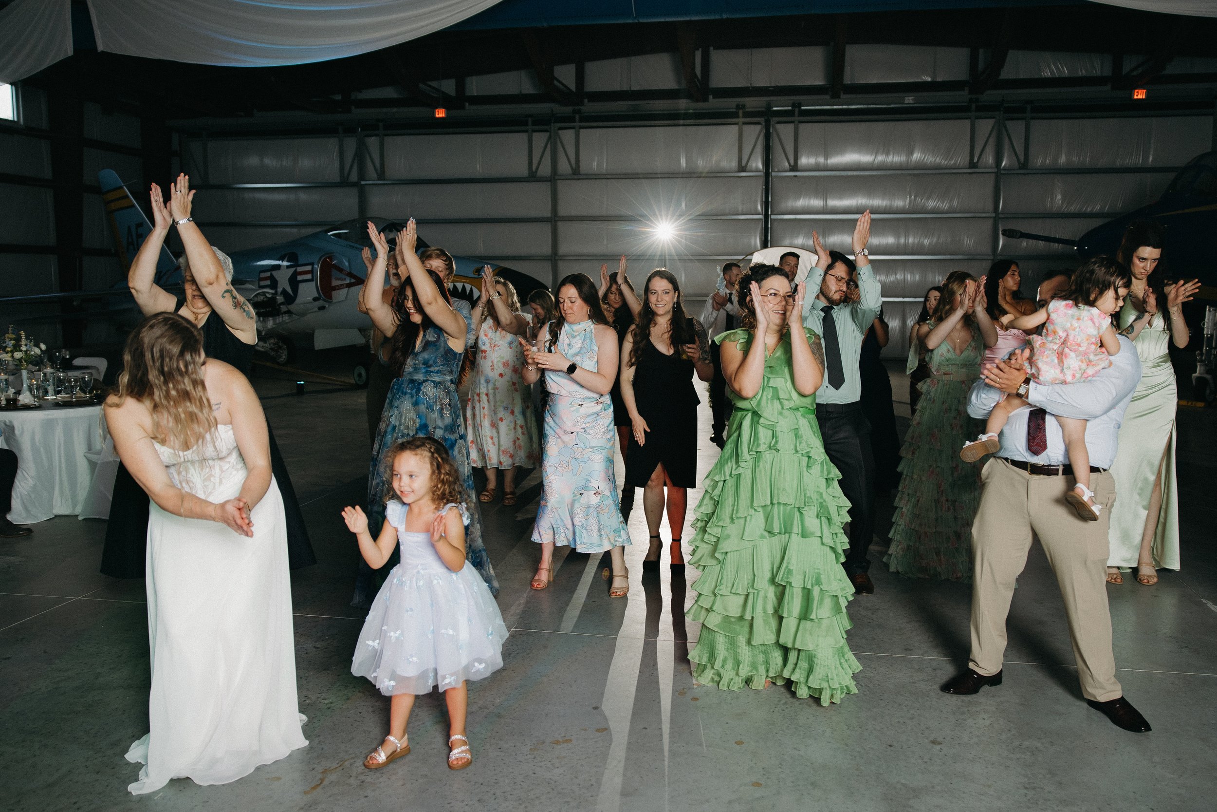 A group of people dancing and celebrating in an indoor hangar with vintage aircraft planes in the background.