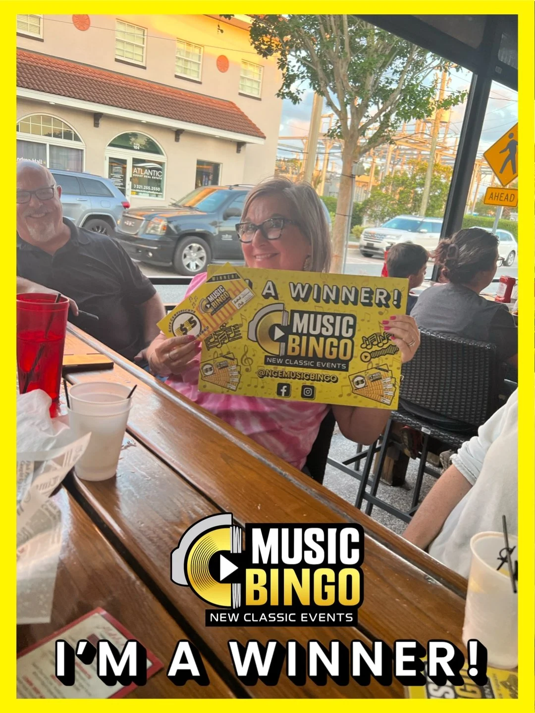 Woman holding a yellow sign that says "A WINNER! MUSIC BINGO" inside a restaurant, smiling, with other people seated at tables nearby, and cars visible through the window.