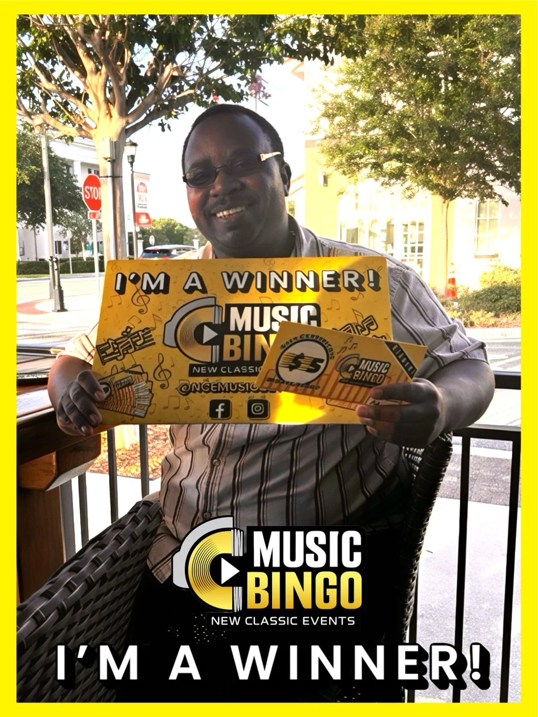 A man smiling and holding a yellow sign that reads 'I'm a winner! Music Bingo.' He is sitting outside on a patio with trees and buildings in the background. The image has a yellow border with a black and yellow logo at the bottom and white text that 
