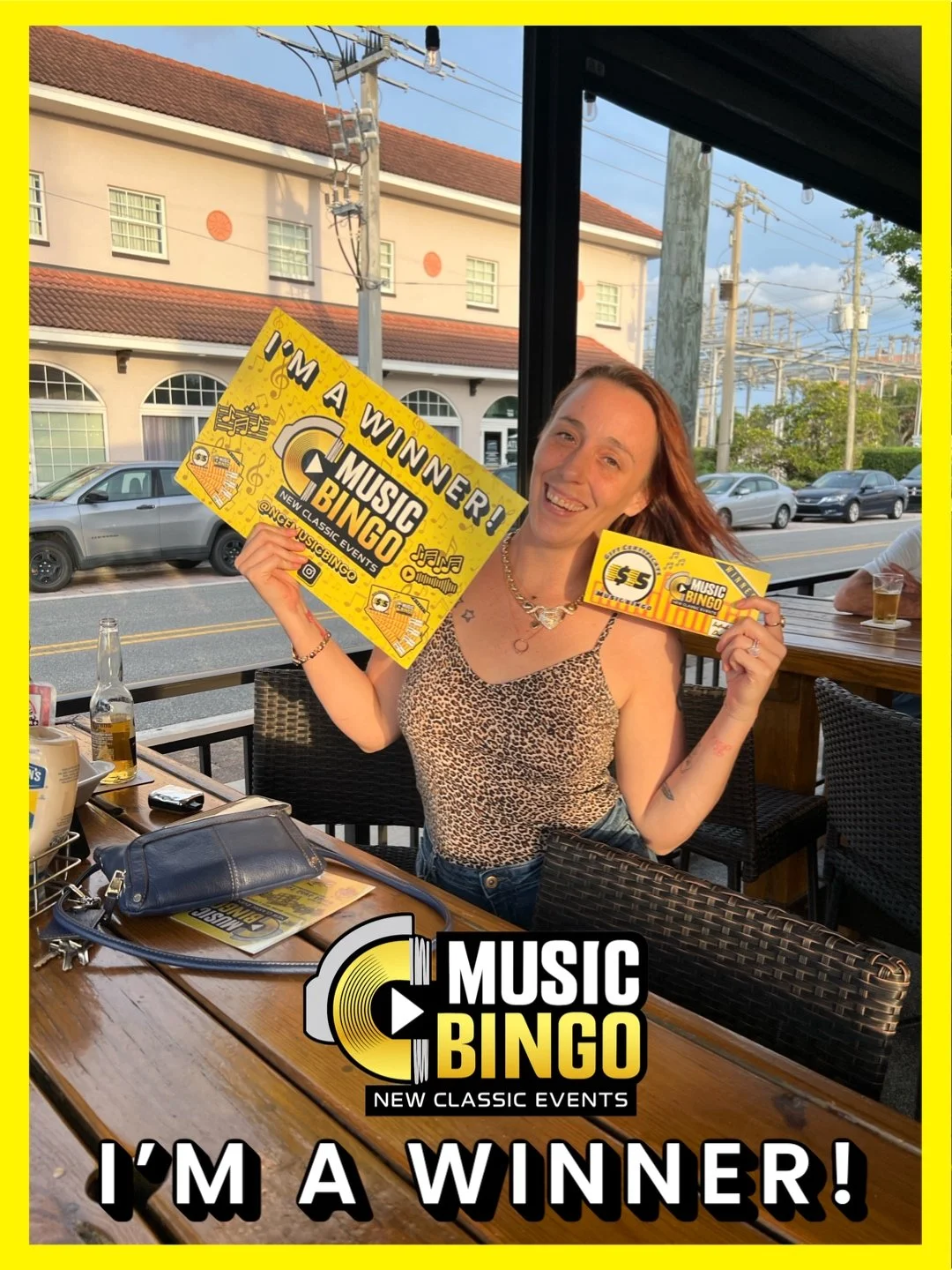A woman with red hair smiling and holding up a large yellow sign that reads 'I'm a Winner! Music Bingo' and a smaller ticket for $5. She is sitting at an outdoor table with drinks and personal items, with a street and building in the background.