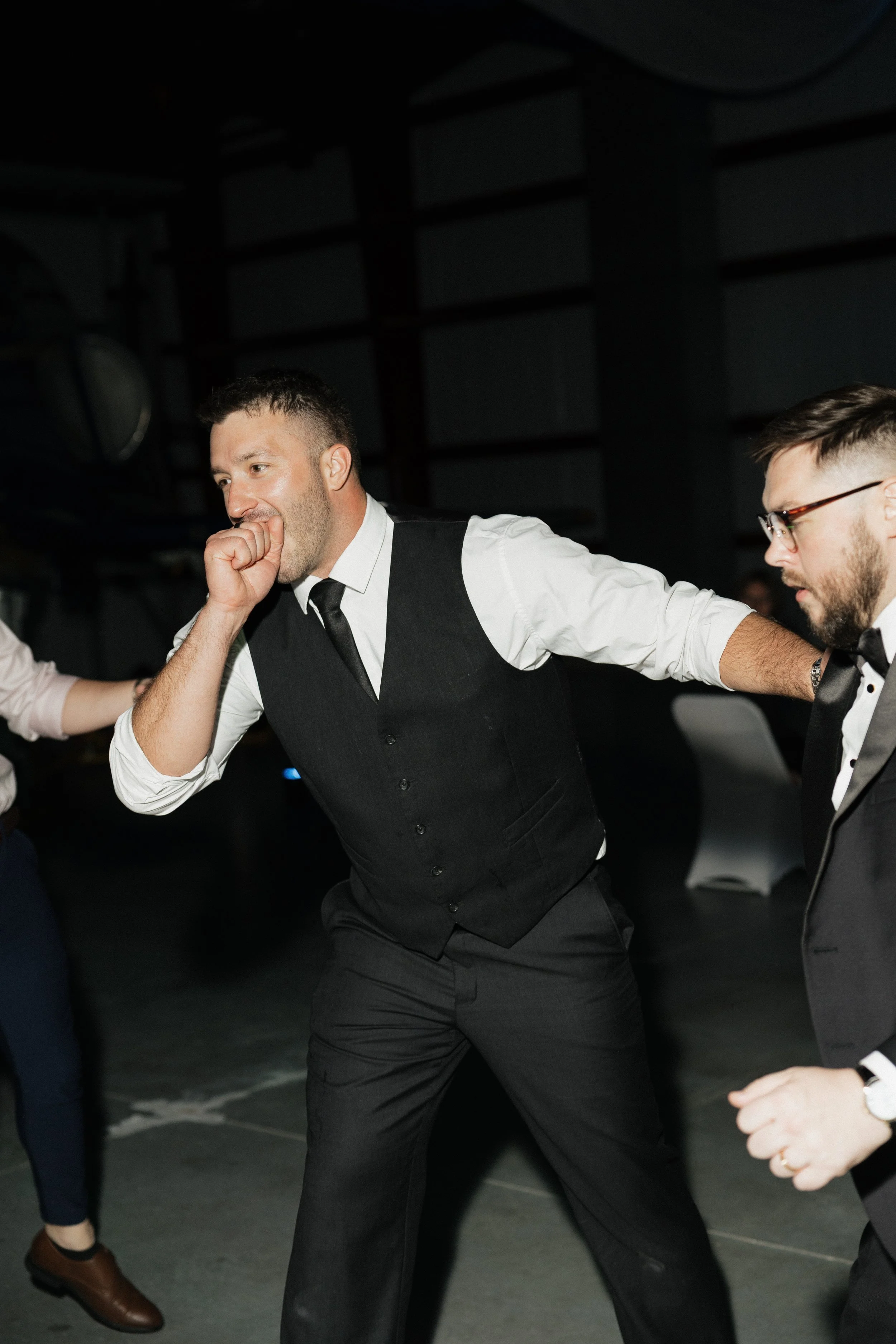 Two men dressed in formal attire are dancing together at an event. One man has short dark hair, a beard, and is wearing a black vest over a white shirt with a black tie. The other man has glasses, a beard, and is wearing a black tuxedo. They are hold