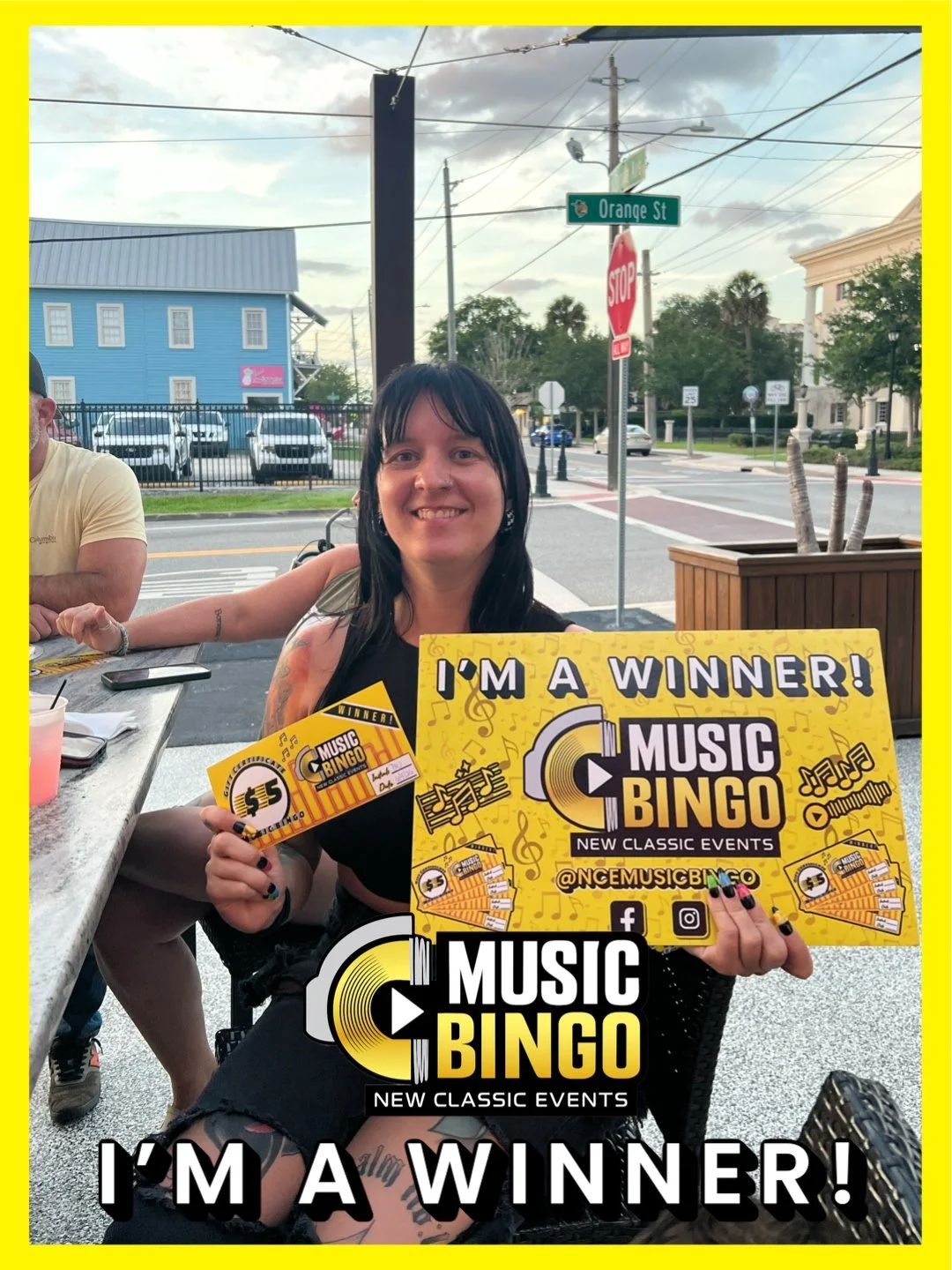 A woman with black hair, tattoos, and colorful nails smiling and holding yellow bingo tickets with the words 'I'm a winner!' at an outdoor table. She is celebrating her win at a music bingo event.