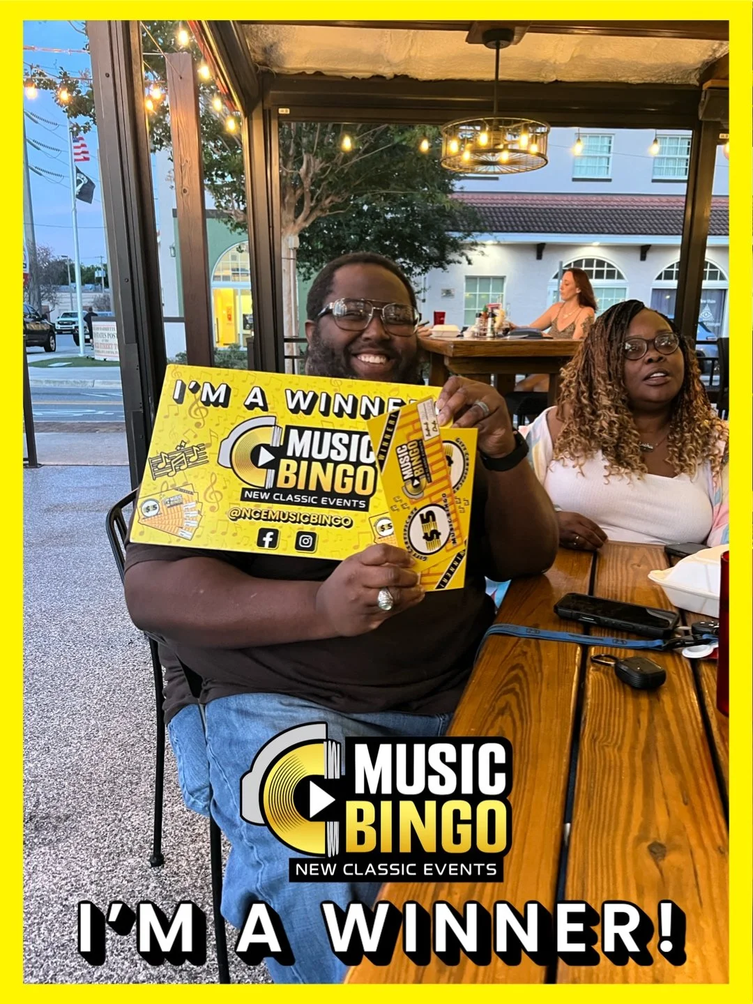 A man smiling and holding a yellow sign that says 'I'm a Winner! Music Bingo' with a black and yellow logo, sitting at a wooden table with a woman seated beside him inside a restaurant or cafe with string lights and large windows in the background.