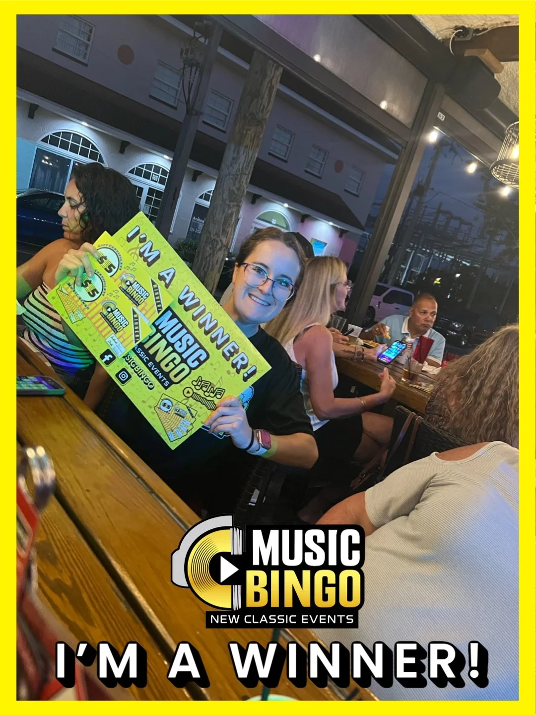 A woman is smiling and holding a sign that says 'I'm a Winner!' at a music bingo event in a restaurant or bar with several other people seated at tables, some using phones, in a warmly lit environment with large windows showing a street scene outside