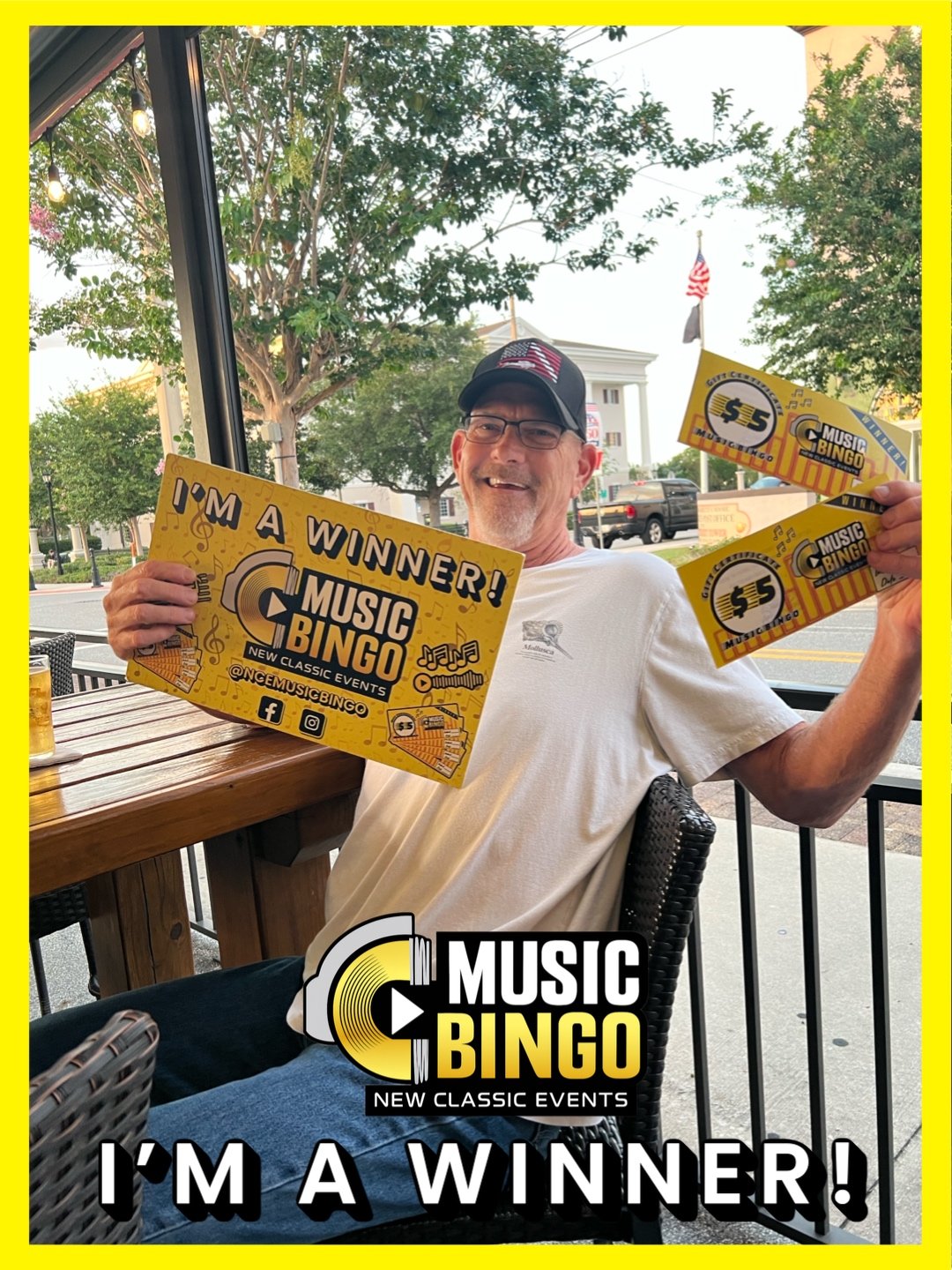 A man wearing glasses and a baseball cap holding four yellow music bingo tickets with the message 'I'm a winner!' while sitting at a table inside a restaurant or cafe. Outside visible through the window are trees, a street, and buildings.
