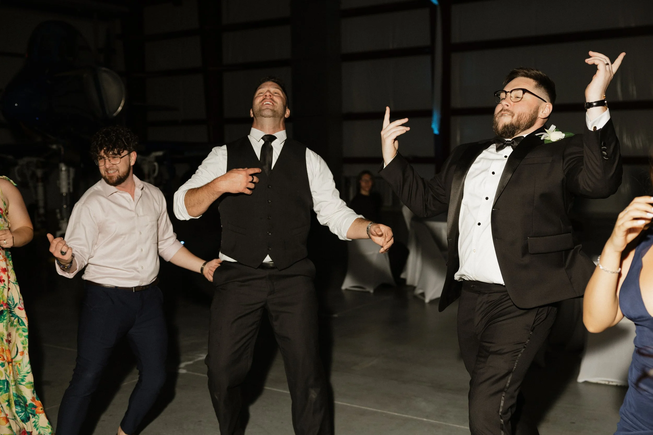 Group of men dancing at a celebration or party, some wearing formal attire like tuxedos, in an indoor setting.