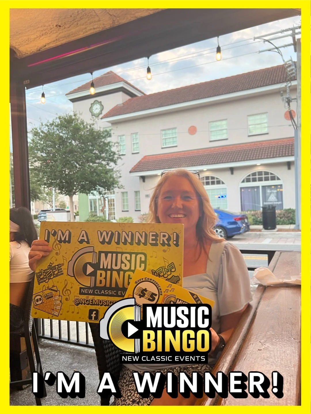 Woman smiling and holding a yellow sign that says 'I'm a winner!' at a music bingo event, with a logo reading 'Music Bingo New Classic Events' on the photo.
