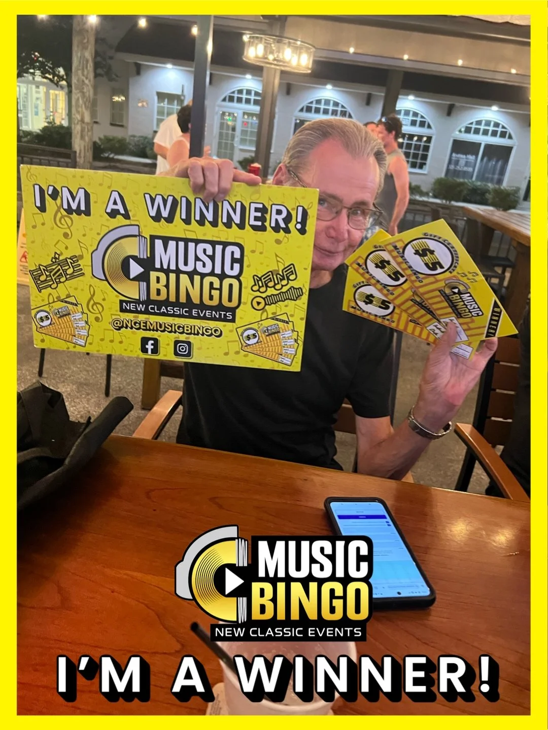 A man sitting at a table holding up a yellow sign that reads 'I'm a winner! Music Bingo' with social media icons and a logo for Music Bingo. He is also holding several yellow flyers or tickets with the same design. The setting appears to be an outdoo