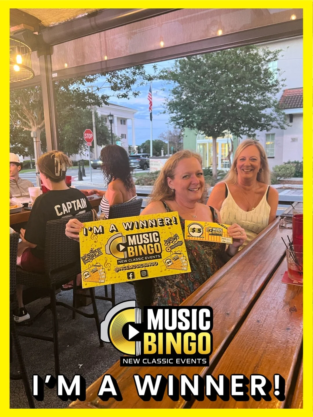 Two women smiling and holding music bingo winning signs at a restaurant or bar, with other patrons sitting at tables, large windows, trees, and buildings outside in the background.