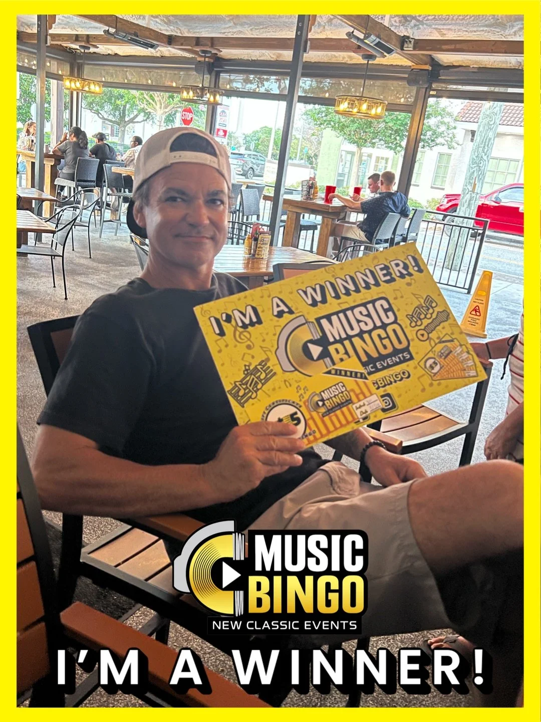 A man sitting in a restaurant holding a yellow sign that says 'I'm a winner! Music Bingo'. He is wearing a black t-shirt and a white cap backward. The restaurant has large windows, many tables, and people dining.