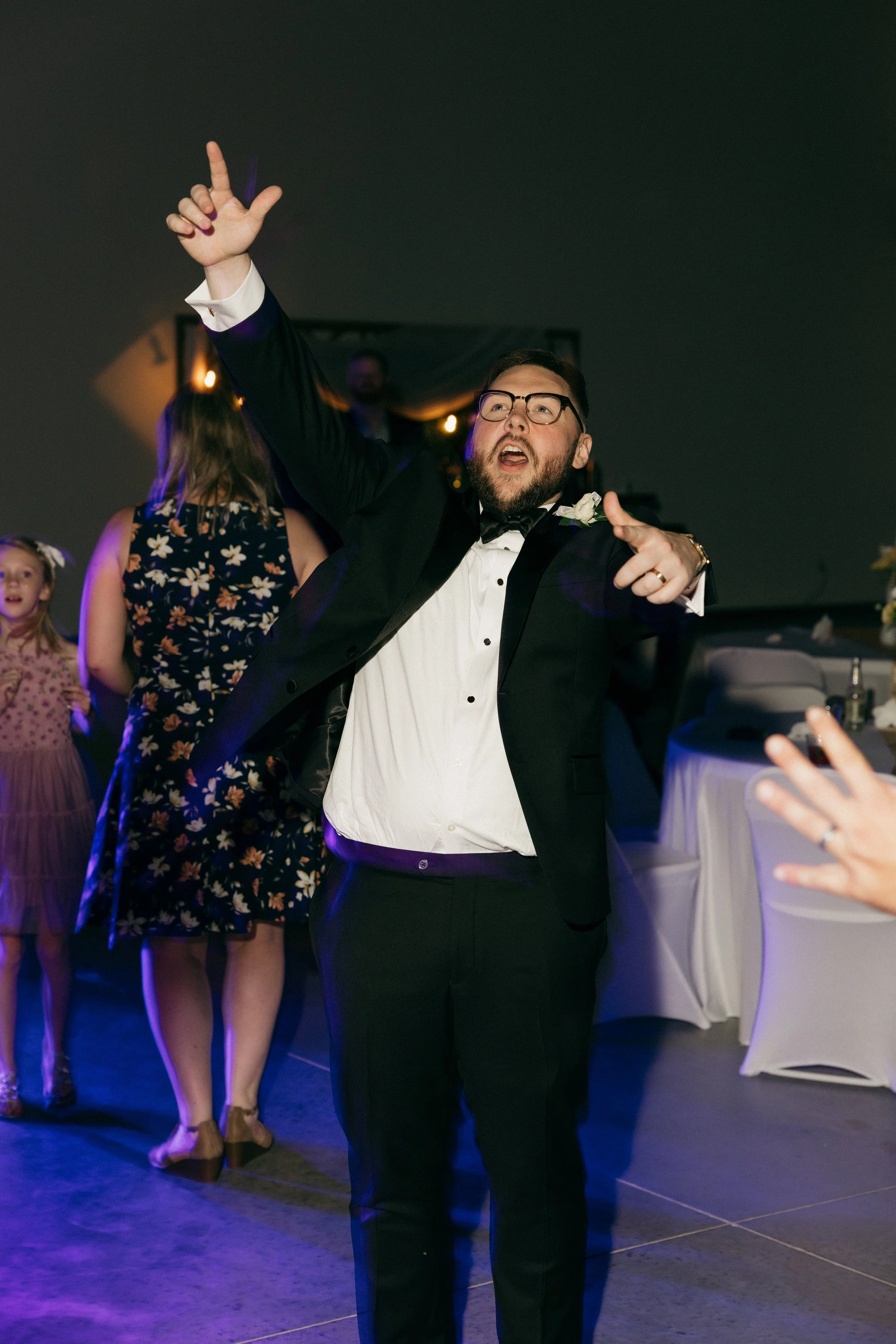 A man in a tuxedo with glasses dancing enthusiastically at a party, with women and children in the background.