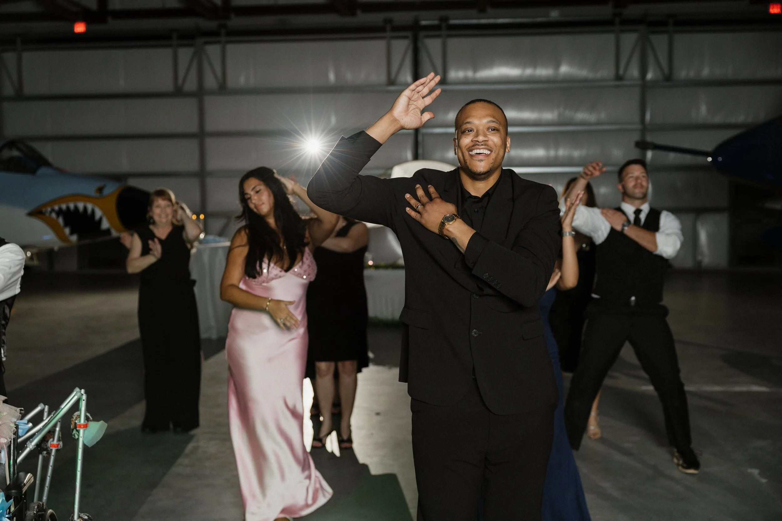 A man in a black suit smiling and waving at an indoor event surrounded by people in formal attire.