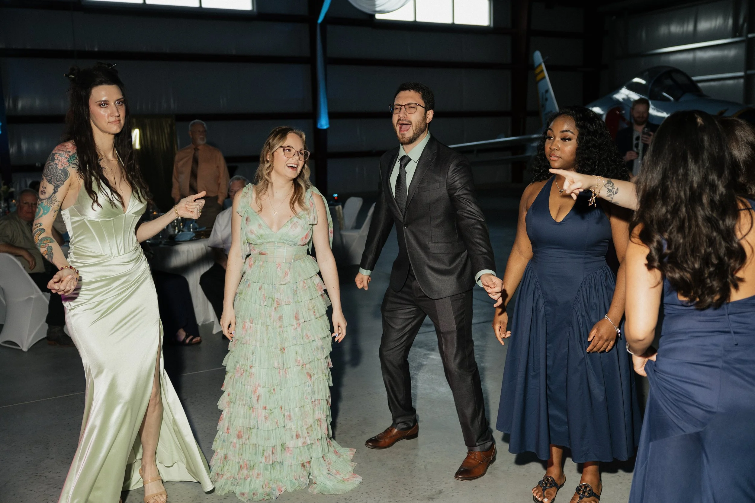 Group of women and a man dancing and talking at an indoor event, with a small airplane in the background.