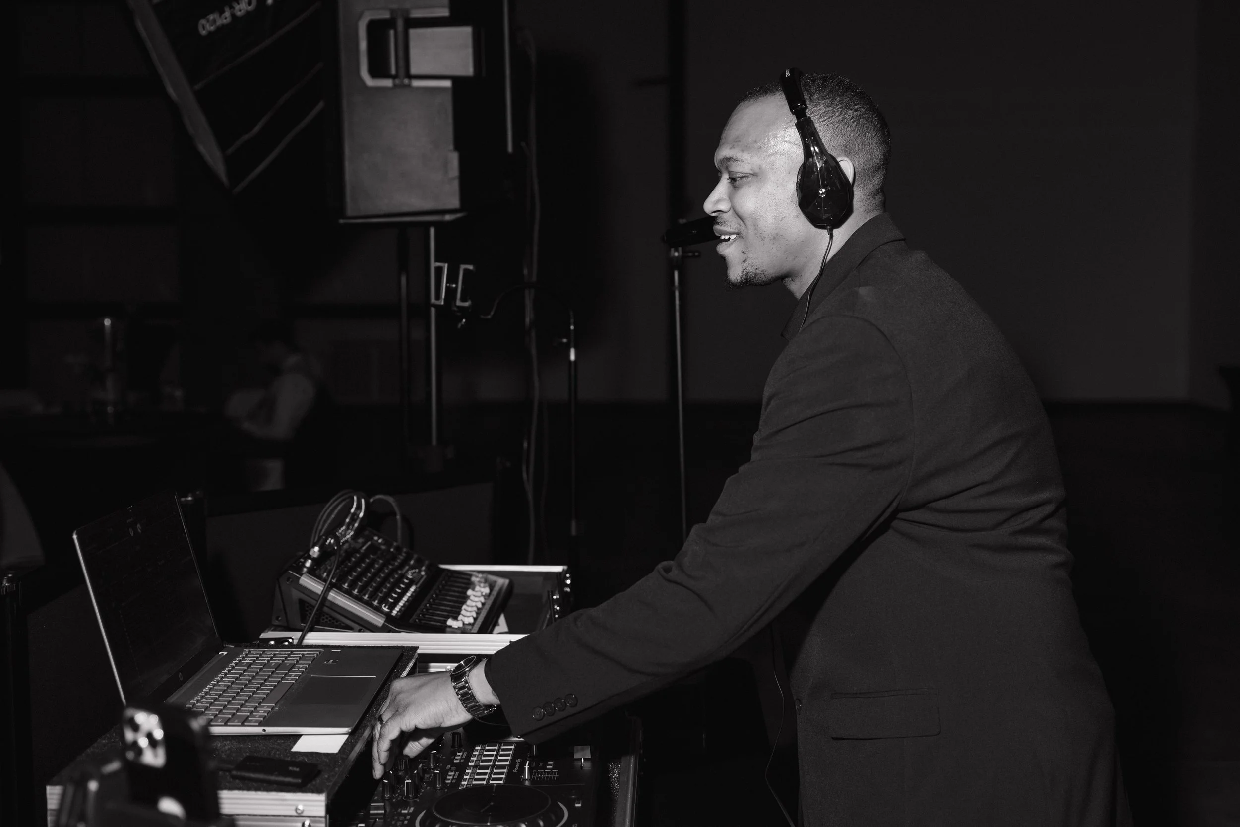 A man in a suit wearing headphones is operating DJ equipment, including a laptop, mixer, and turntable, at a dark indoor setting.