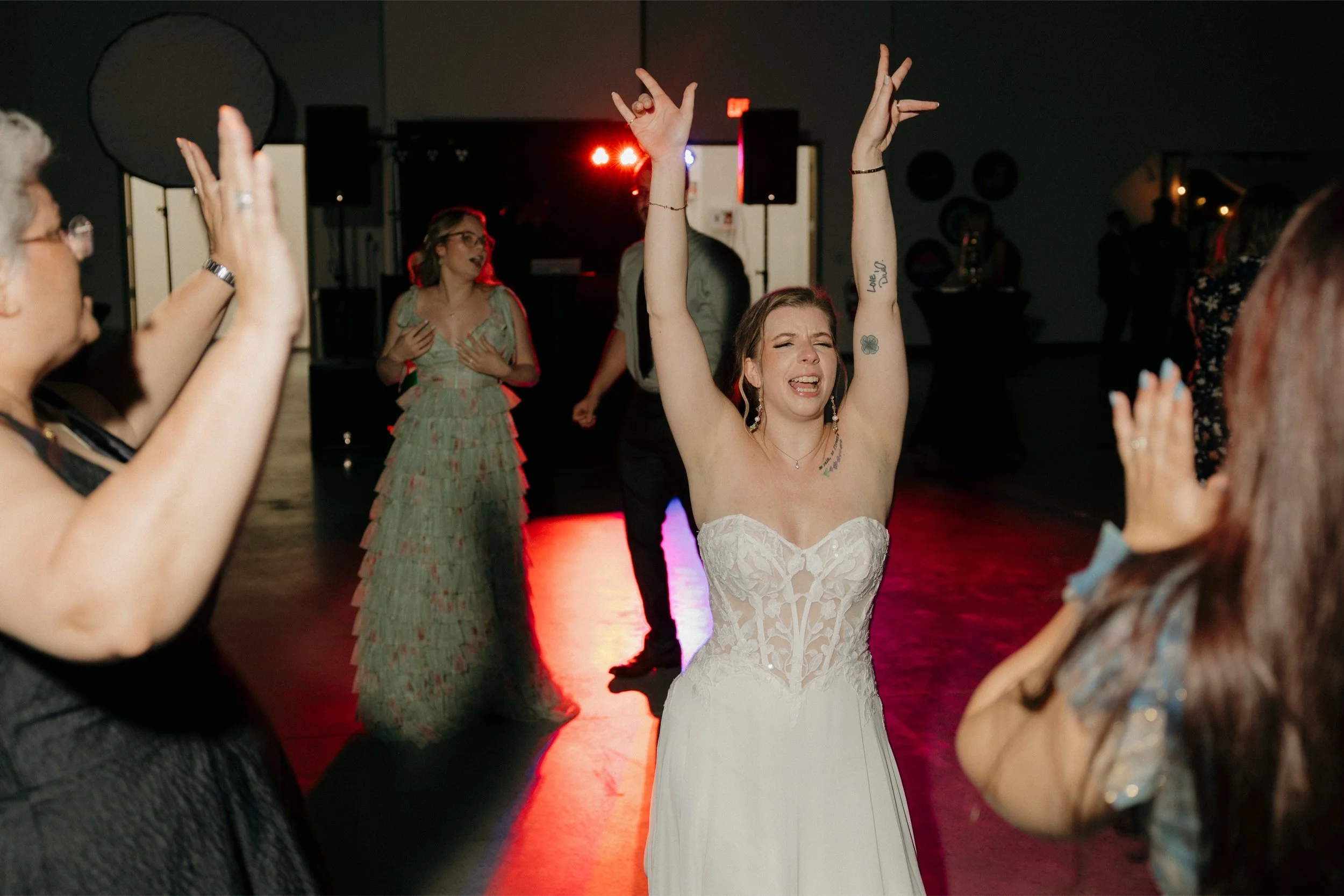 A woman in a strapless white dress with open eyes and a joyful expression dances with her arms raised, surrounded by other women at a celebration or wedding reception in a dimly lit room with colorful lighting.