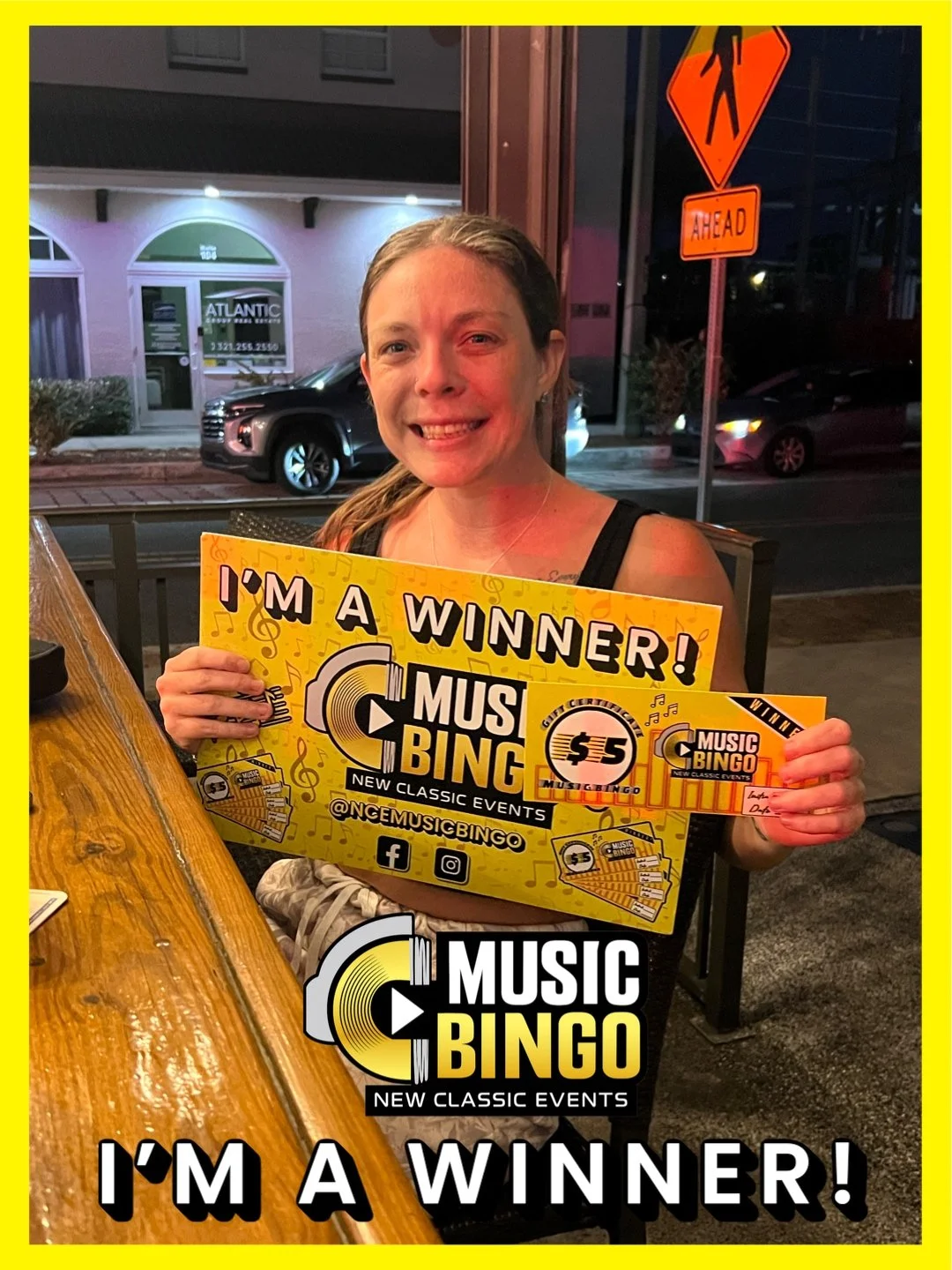 A woman with a smiling face holding a yellow sign that says 'I'm a Winner! Music Bingo, New Classic Events, $5'. She is sitting at a wooden table outside at night, with a crosswalk sign and buildings visible in the background.