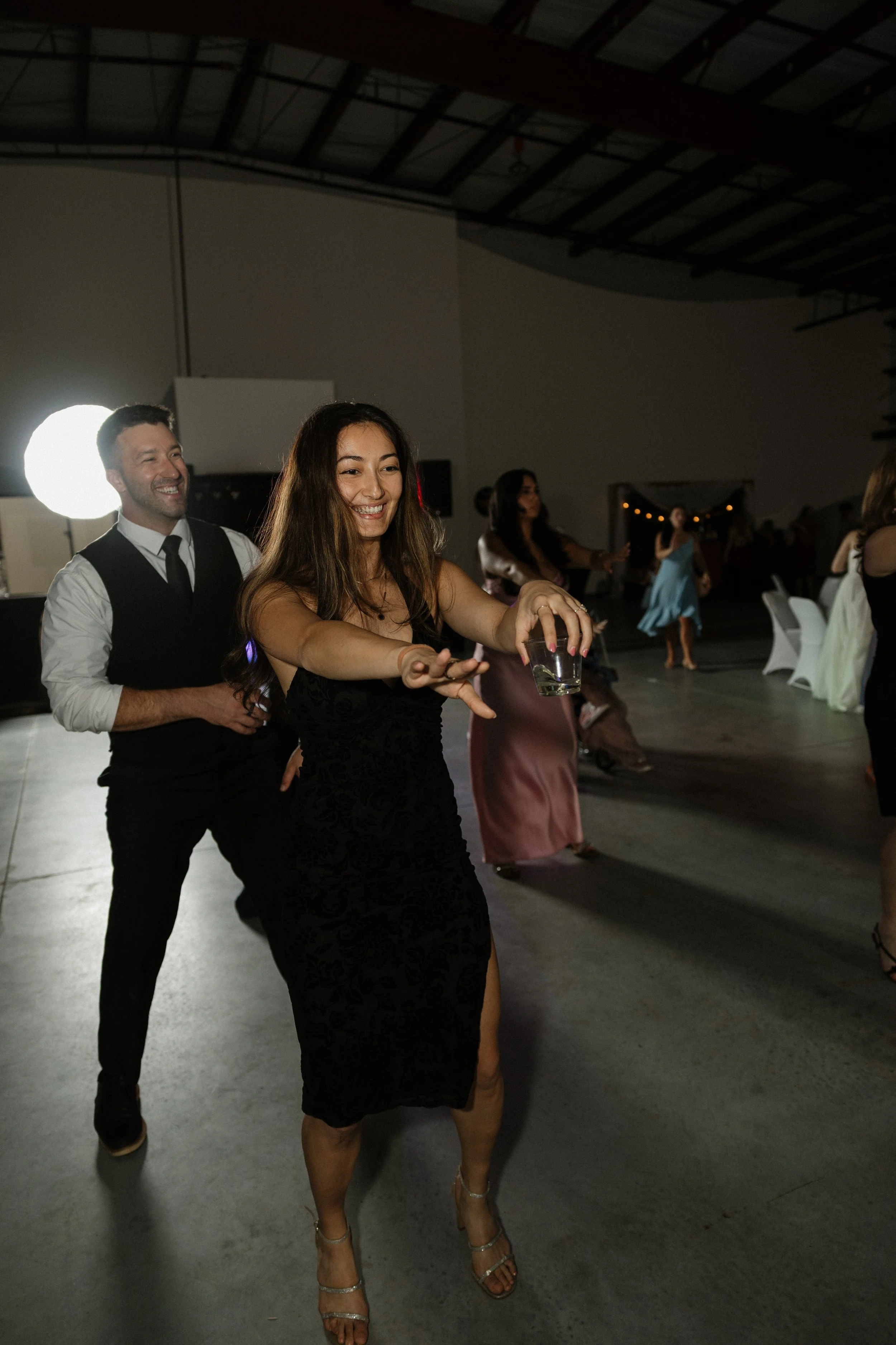 People dancing at a wedding reception under a high ceiling with string lights in the background.