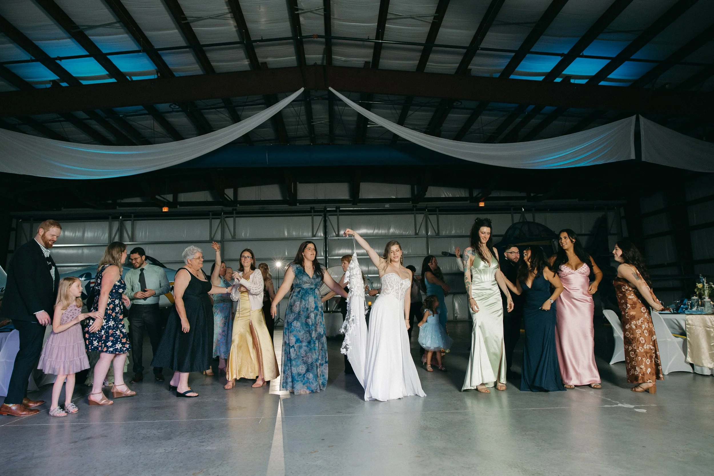 A group of people, including children and adults, dancing and celebrating at a wedding reception inside a large industrial-style venue. The bride is holding a hanger with a wedding dress, and the guests are smiling and having a good time.