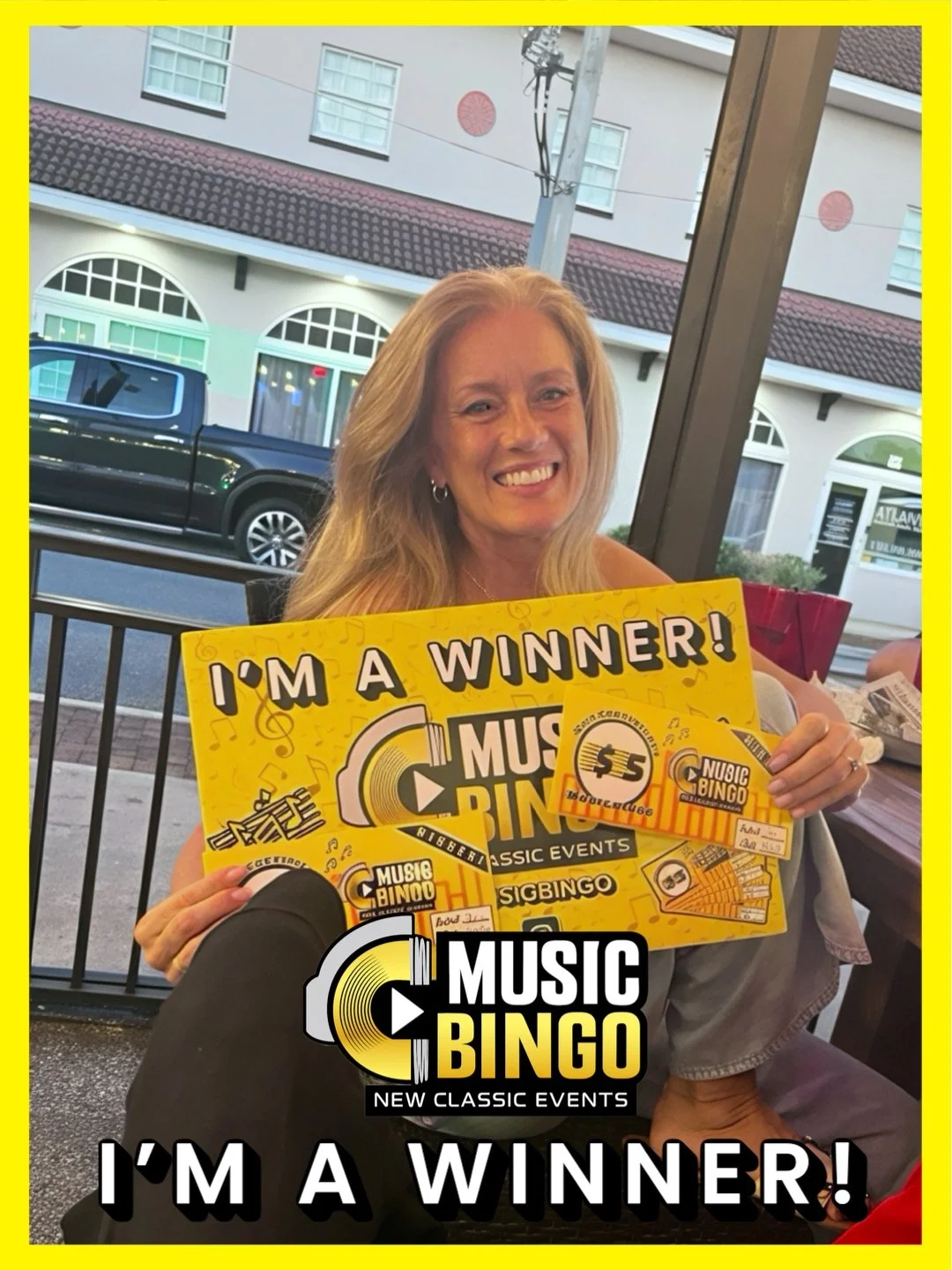 A woman with blonde hair smiling, holding a yellow sign that says 'I'm a Winner!'. She is at a table with other items, and there are buildings and a car outside the window.