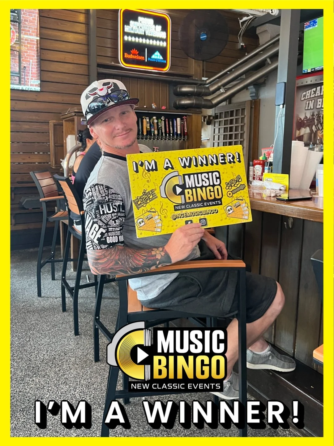 A man sitting at a bar holding a bright yellow sign that reads 'I'm a Winner! Music Bingo, New Classic Events.' He is wearing a gray T-shirt, black shorts, sunglasses on his cap, and has tattoos on his arm. The background shows a wooden wall, a beer 
