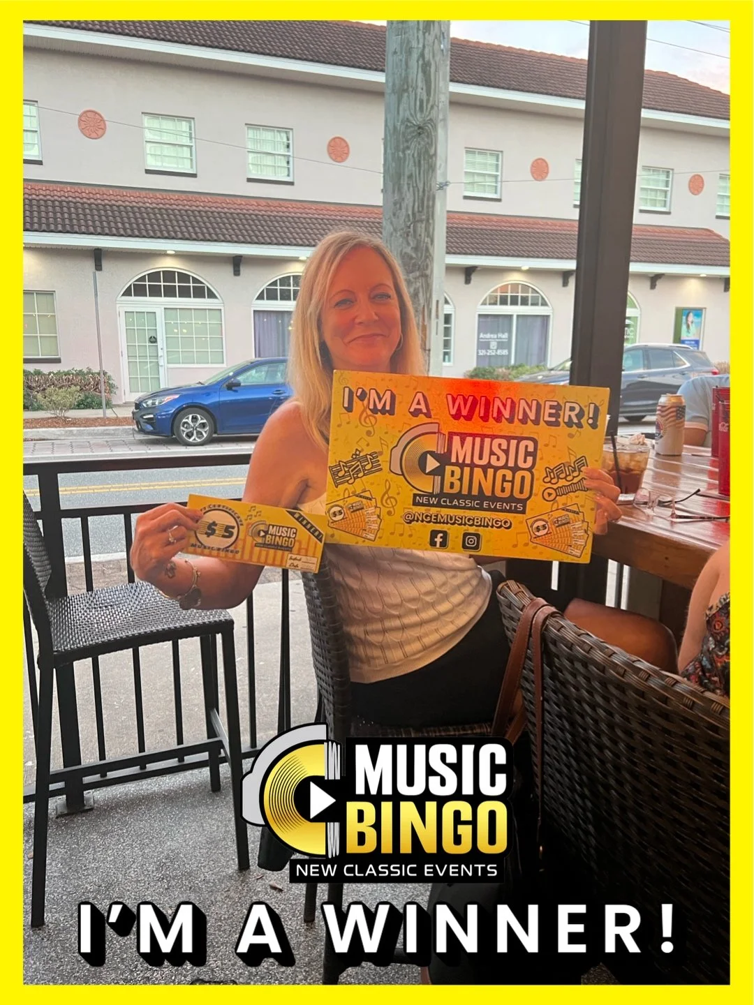 A woman smiling and holding a large Music Bingo winner sign and a $5 music bingo ticket, seated at an outdoor restaurant or cafe with cars and buildings visible in the background.