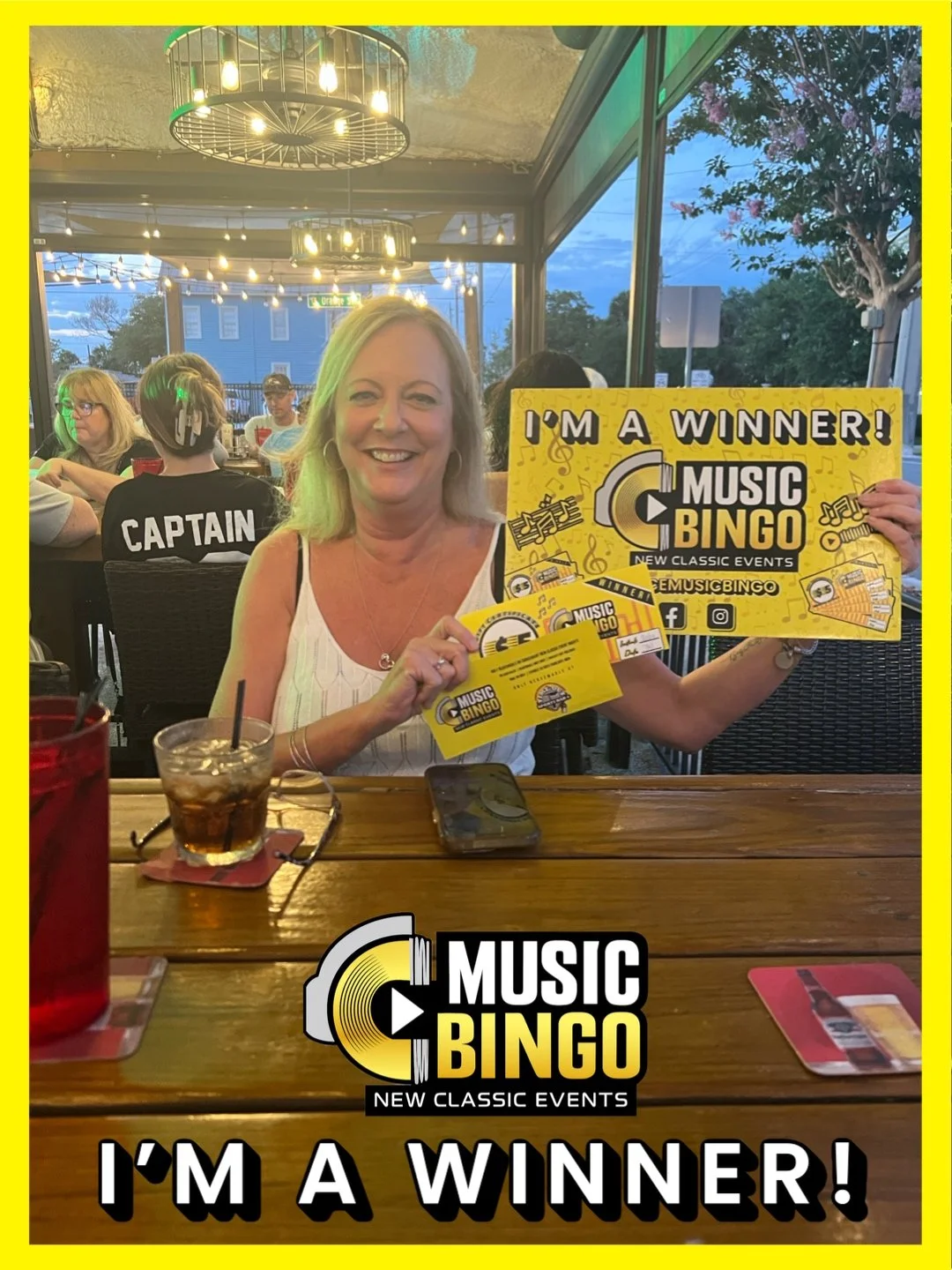 A woman smiling and holding yellow Music Bingo victory cards at a restaurant or bar, with a sign indicating she is a winner, surrounded by other patrons.