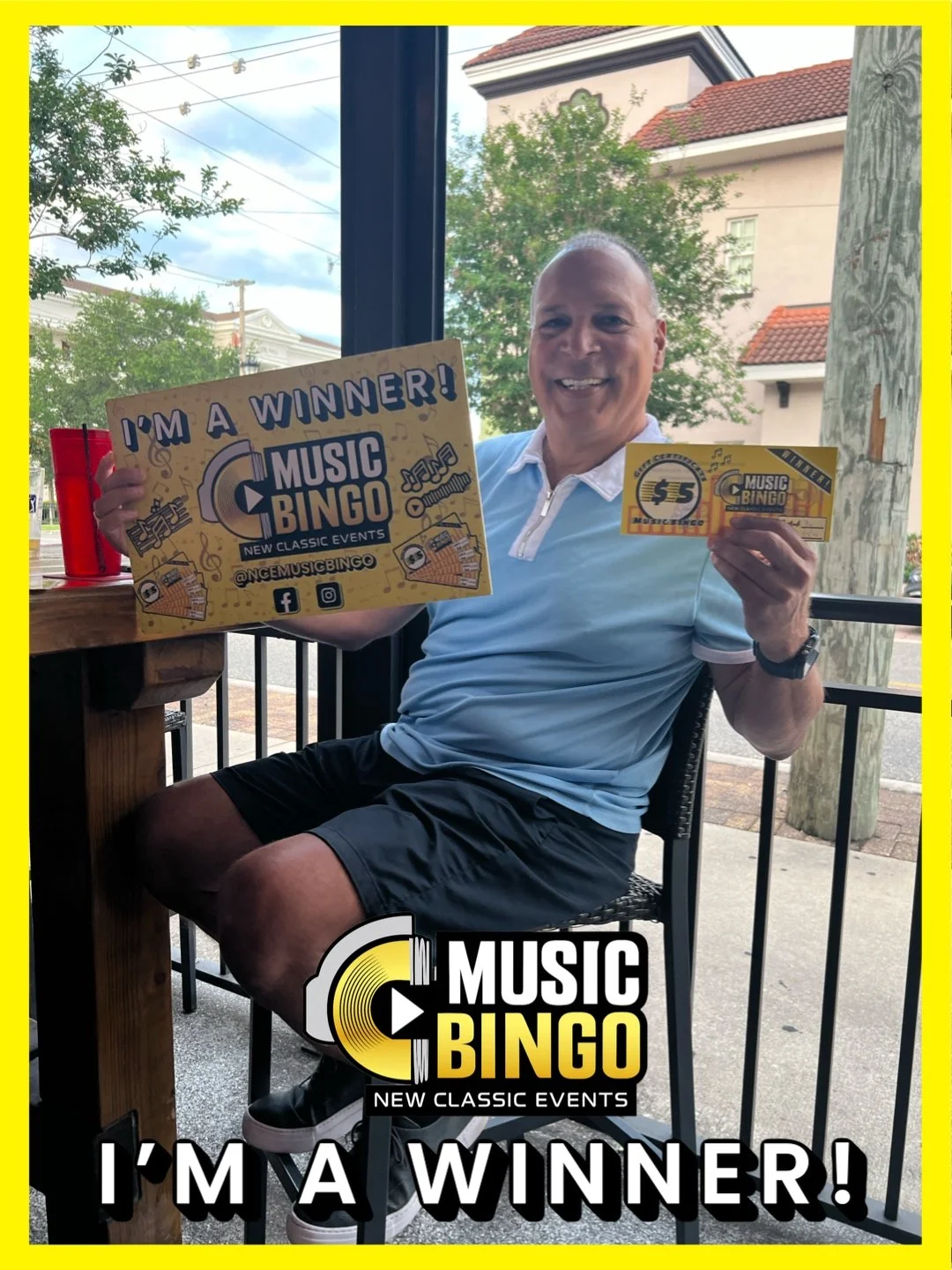 A smiling man sitting at a table holding a sign and tickets for a music bingo event. The sign reads 'I'M A WINNER! MUSIC BINGO.' Outside view through the window shows trees and buildings.