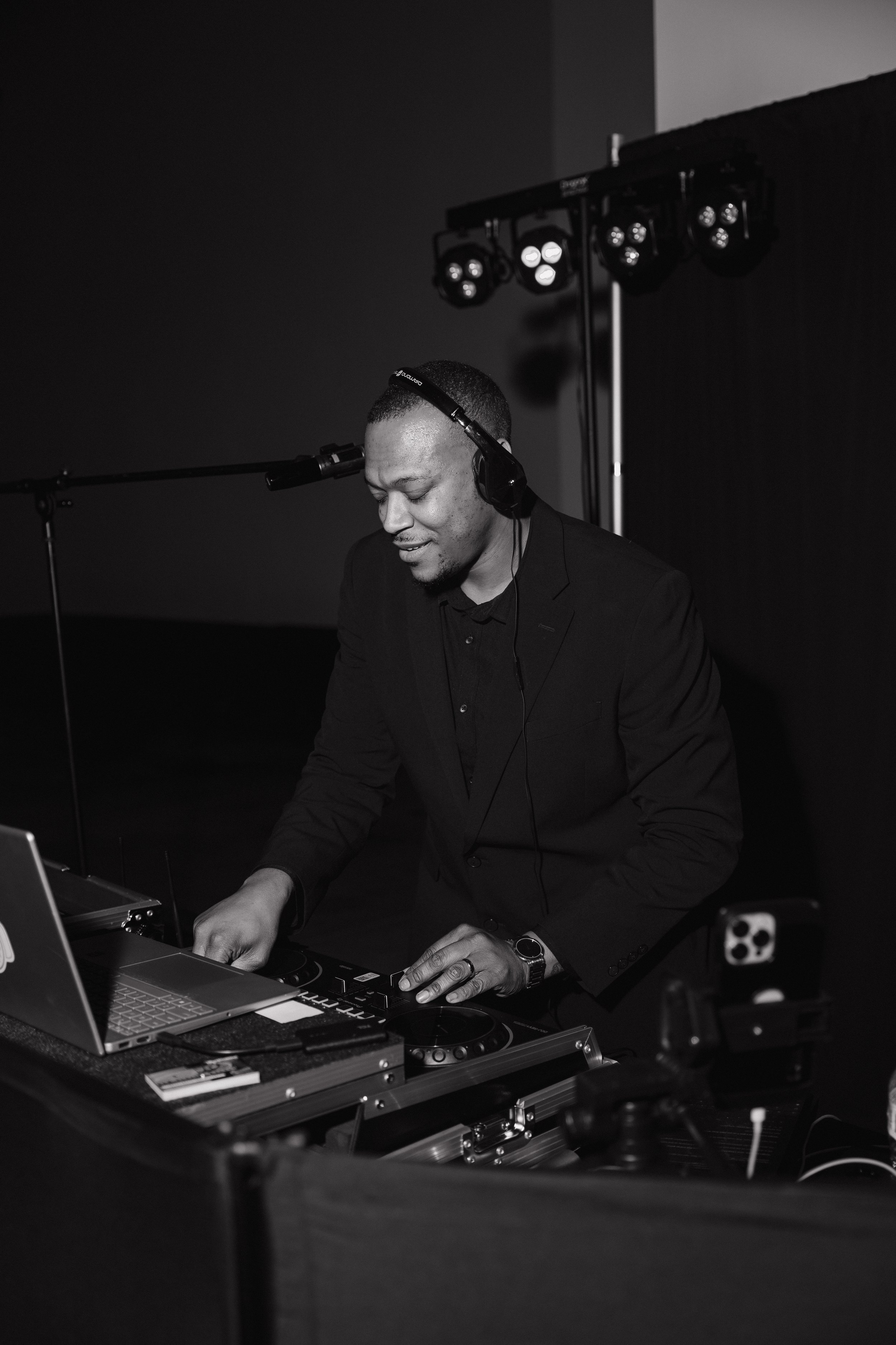 Black and white photograph of a man wearing headphones, playing a keyboard, with a laptop in front of him. Studio lighting equipment is visible in the background.