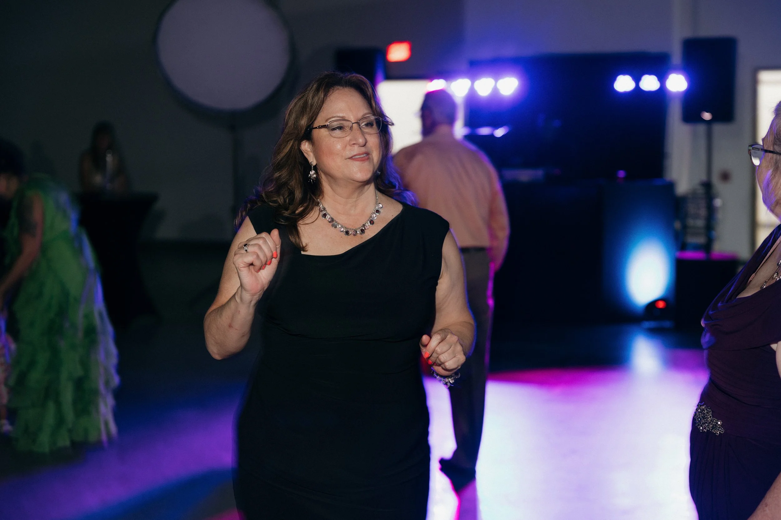 A woman with brown hair, glasses, wearing a black dress and jewelry, dancing at a party with colorful lighting. Other people are visible in the background.