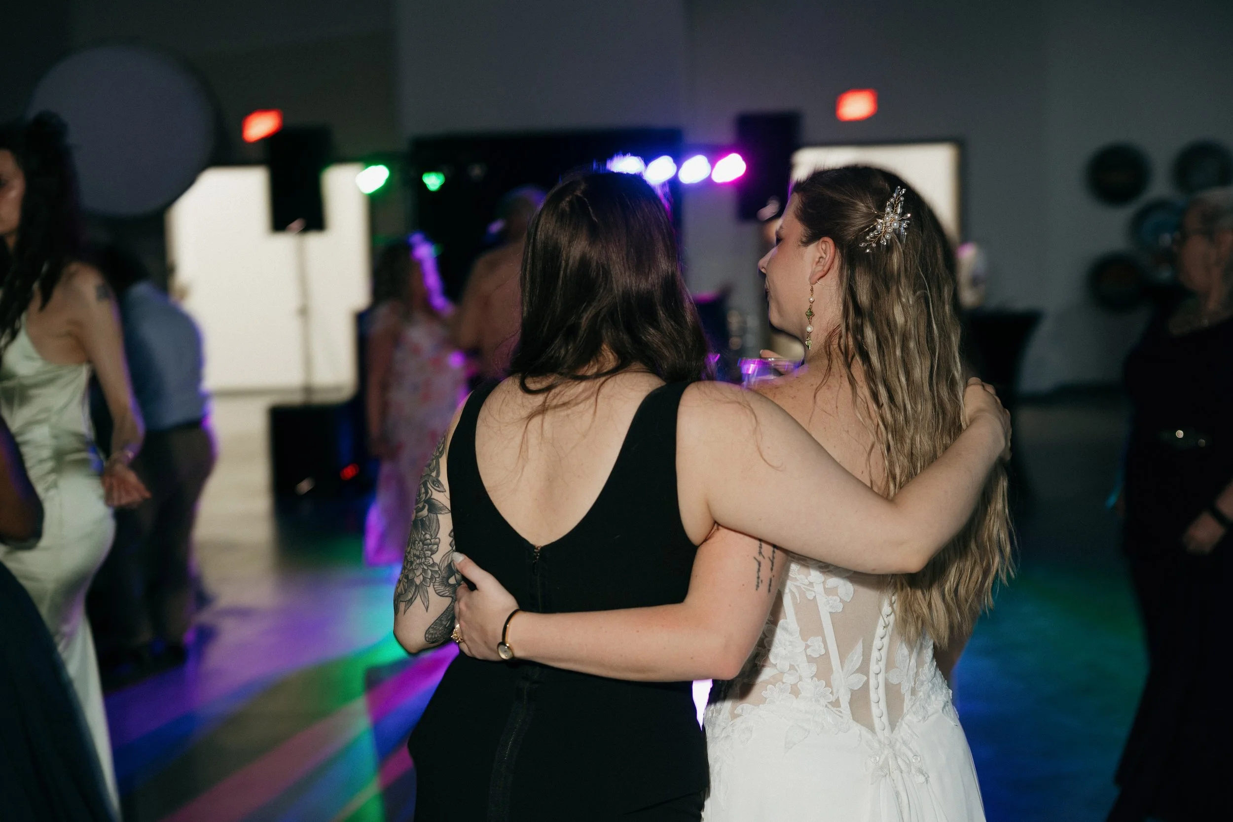 Two women are dancing closely together at a social event, with one wearing a black dress and the other in a white lace dress with a decorative hairpiece.