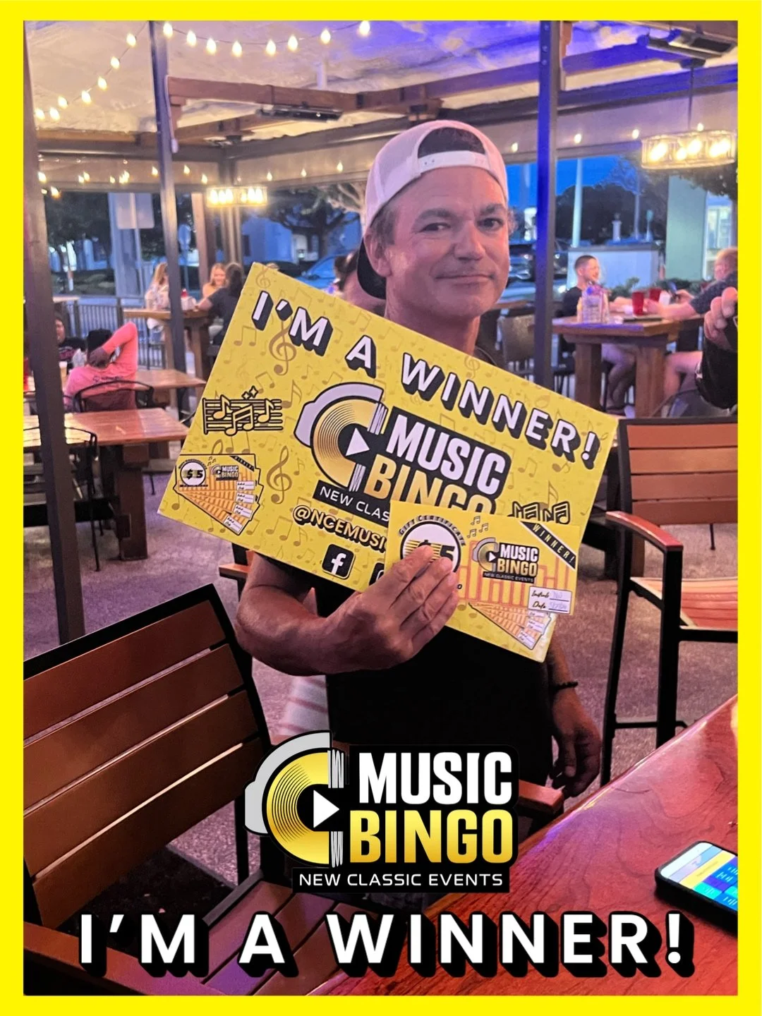 A man wearing a white baseball cap backwards, holding a yellow Music Bingo winner sign at an outdoor event venue with string lights and other people seated at tables.