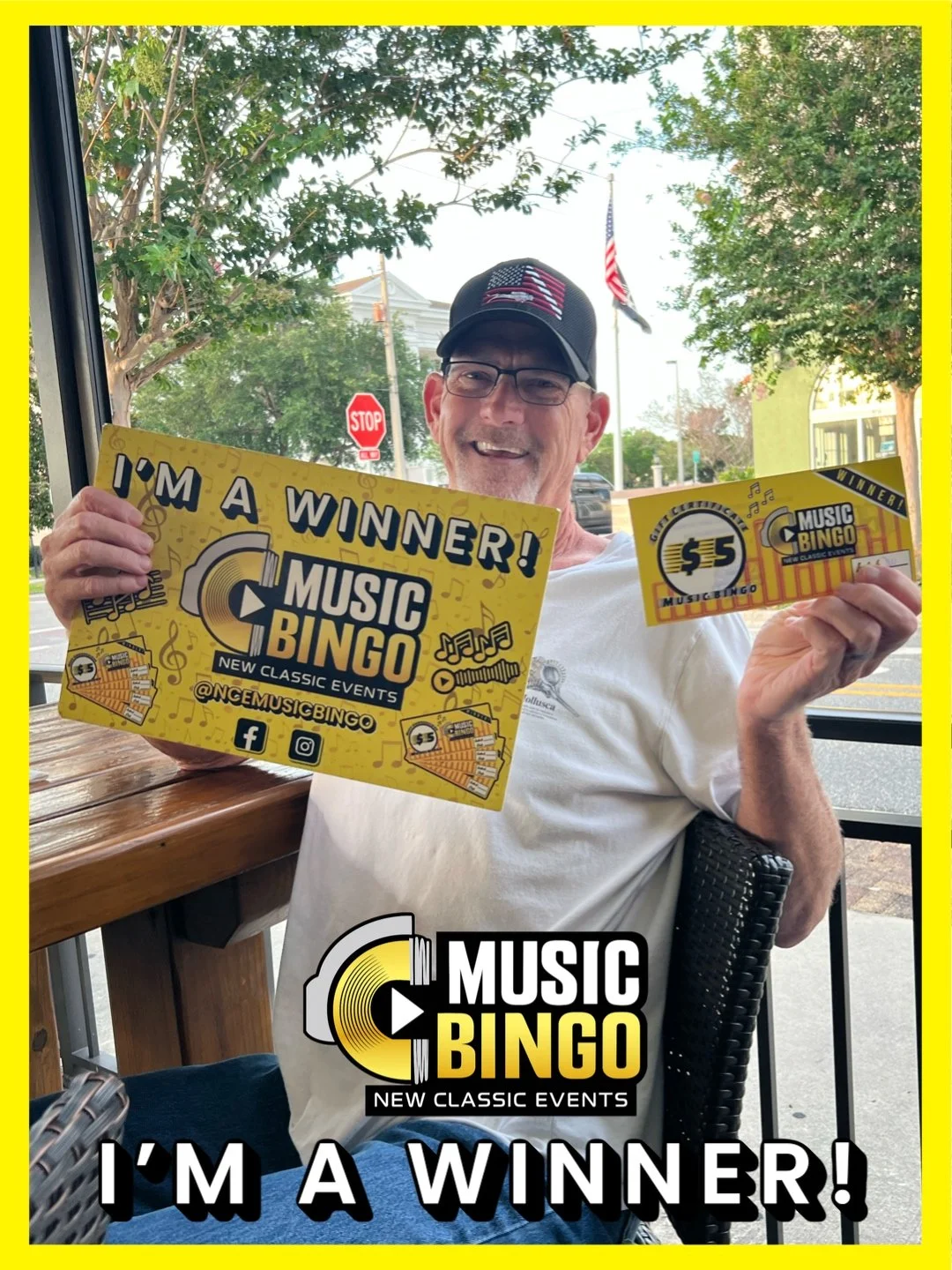 Man sitting at a table inside a restaurant, holding a winner's certificate and a ticket for Music Bingo, smiling. Outside view through window shows trees, American flag, stop sign, and street.