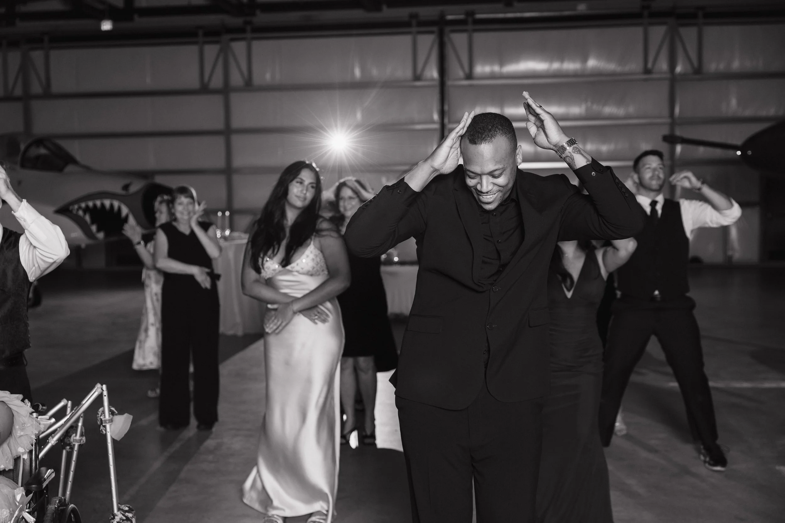 A group of people dancing at a party in an indoor venue, with a man in the foreground smiling and touching his head, surrounded by women in elegant dresses and a man in a vest and tie, with an airplane and warehouse ceiling in the background.
