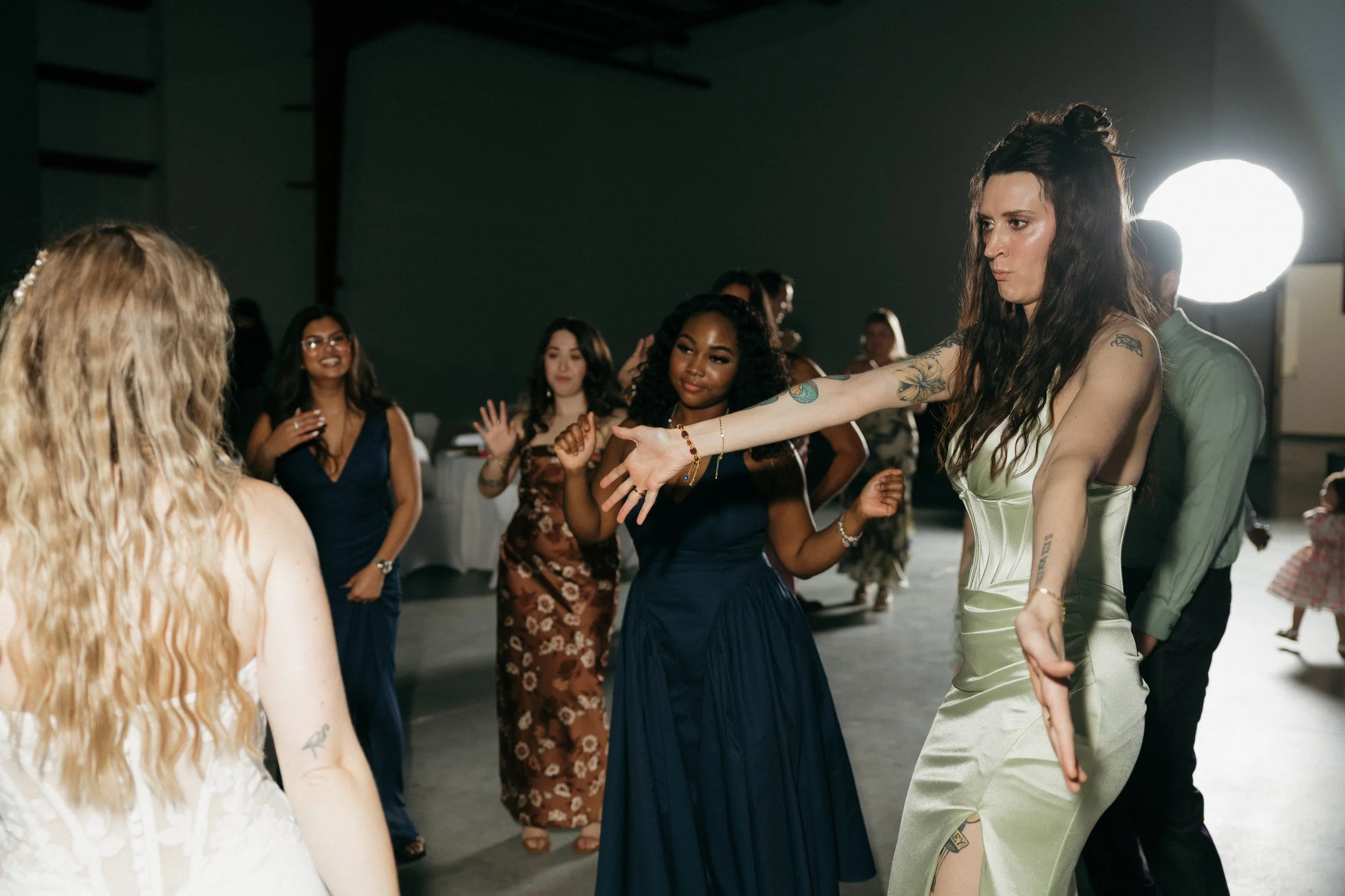 A group of women dancing at an indoor event, with a focus on a woman with long dark hair and tattoos on her arm in the foreground.