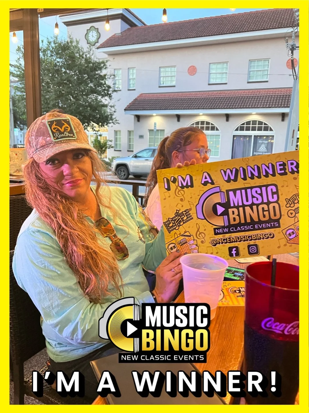Woman with long curly hair wearing a pink cap and sunglasses holding a colorful flyer that reads 'I'm a Winner Music Bingo' at a restaurant table. Another person is seen in the background, and outdoor buildings are visible through the window.