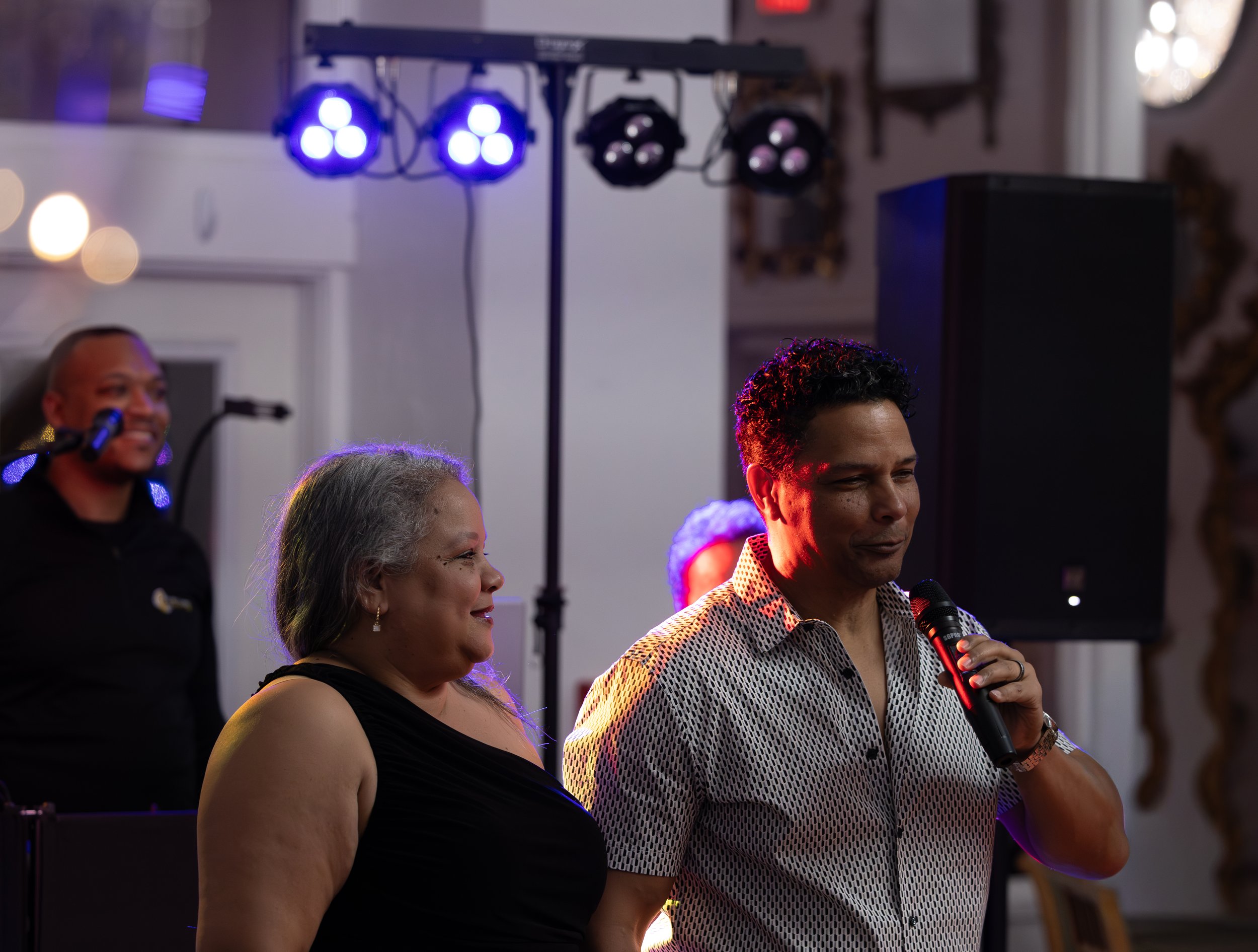 People at an indoor event, with one man holding a microphone, and a woman in a black dress standing beside him. There is DJ equipment and purple lighting in the background.