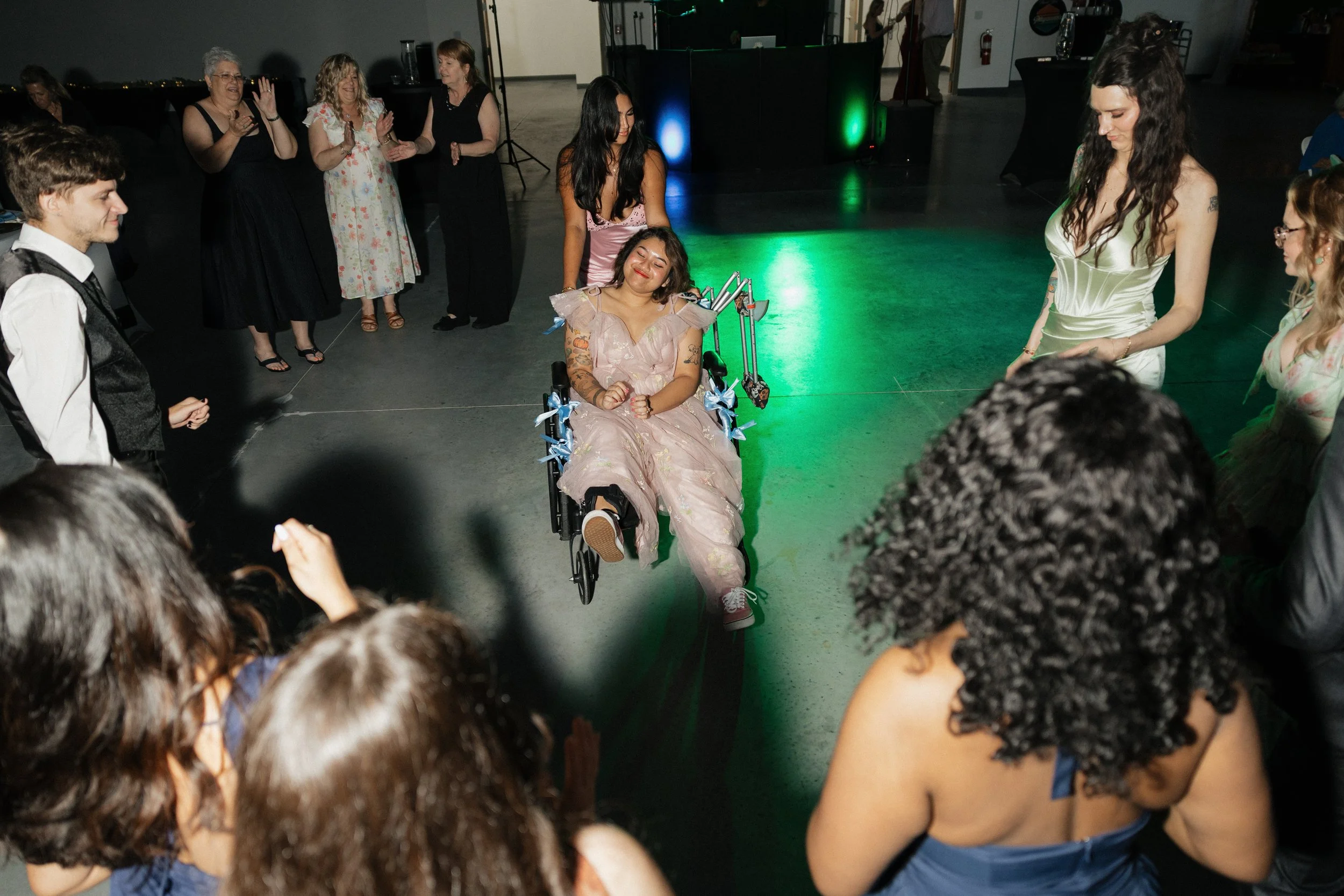 Group of people dancing at an indoor party with colorful lighting, a person in a wheelchair is smiling and dancing in the center.