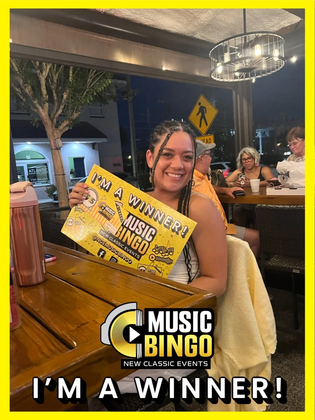 A woman smiling and holding a yellow sign that reads 'I'm a Winner! Music Bingo.' She is sitting at a wooden table in a restaurant or café, with other people in the background and a window showing street outside.