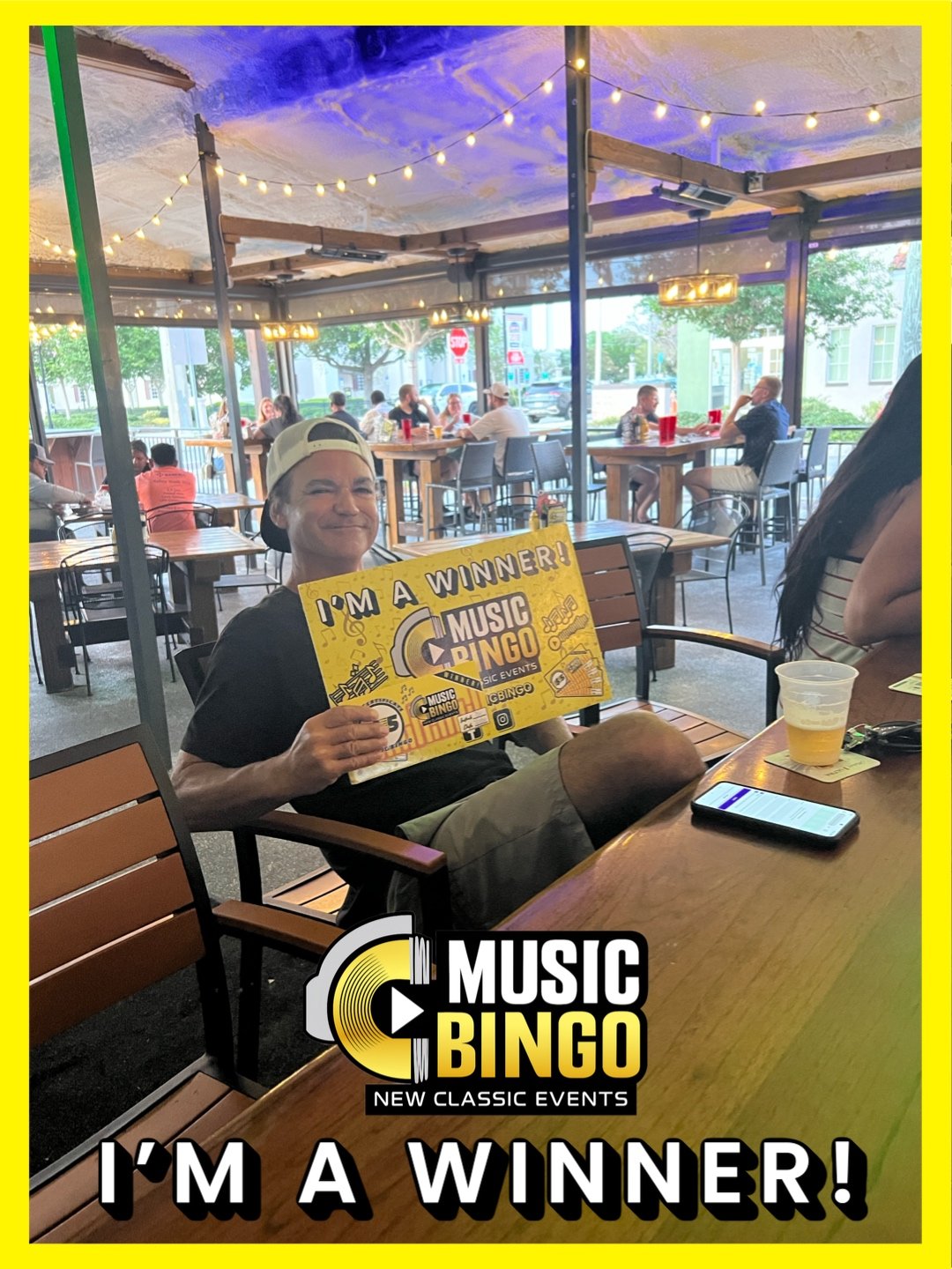 A man sitting at an outdoor restaurant or patio, holding a sign that says "I'm a winner! Music Bingo." He is smiling and wearing a backward baseball cap. There are other people in the background, some sitting at tables and others walking by. The sett