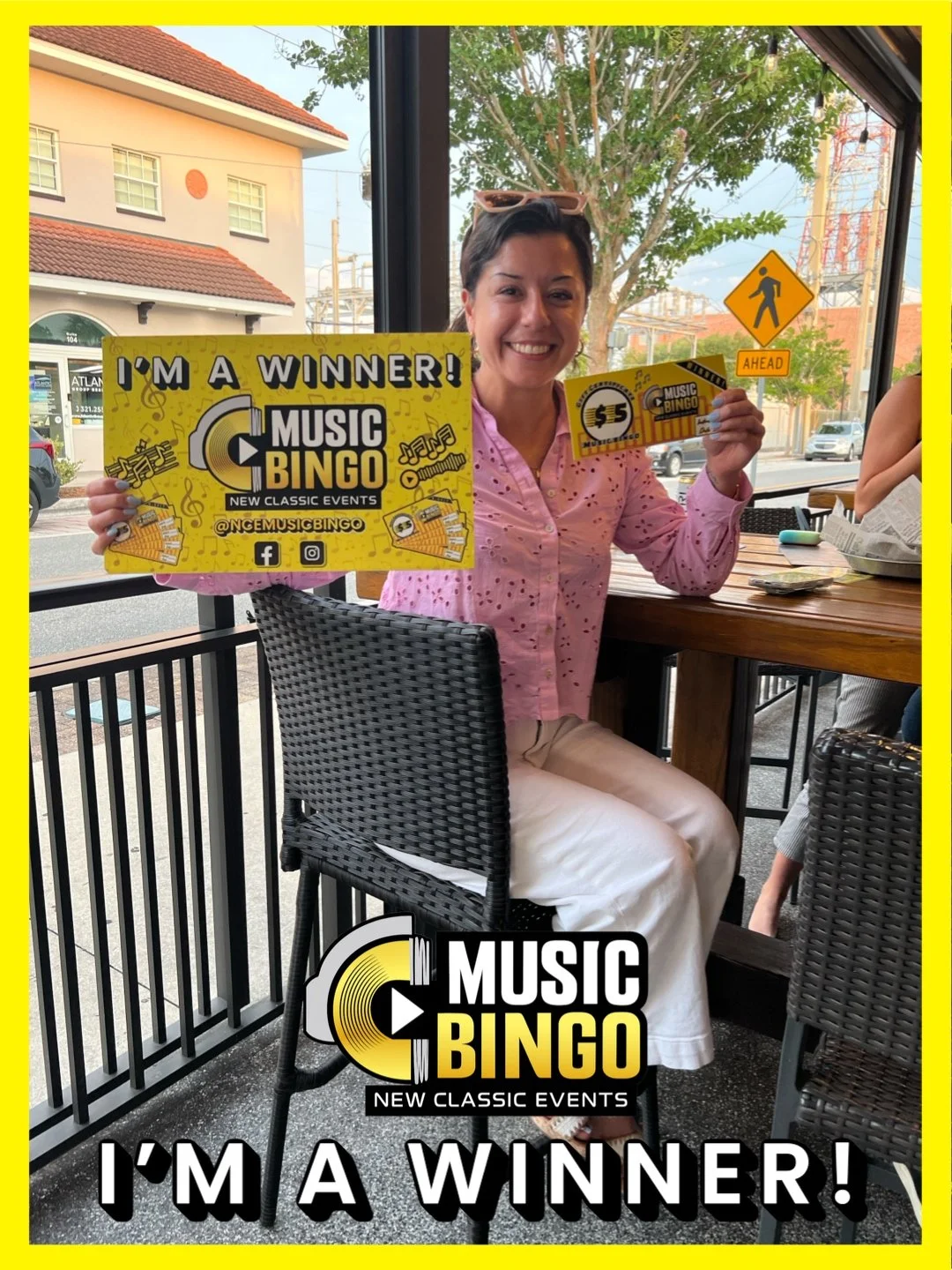 Woman sitting at a restaurant table holding a winning music bingo sign and a $5 prize coupon, smiling at the camera, with a view outside the window of trees and buildings.