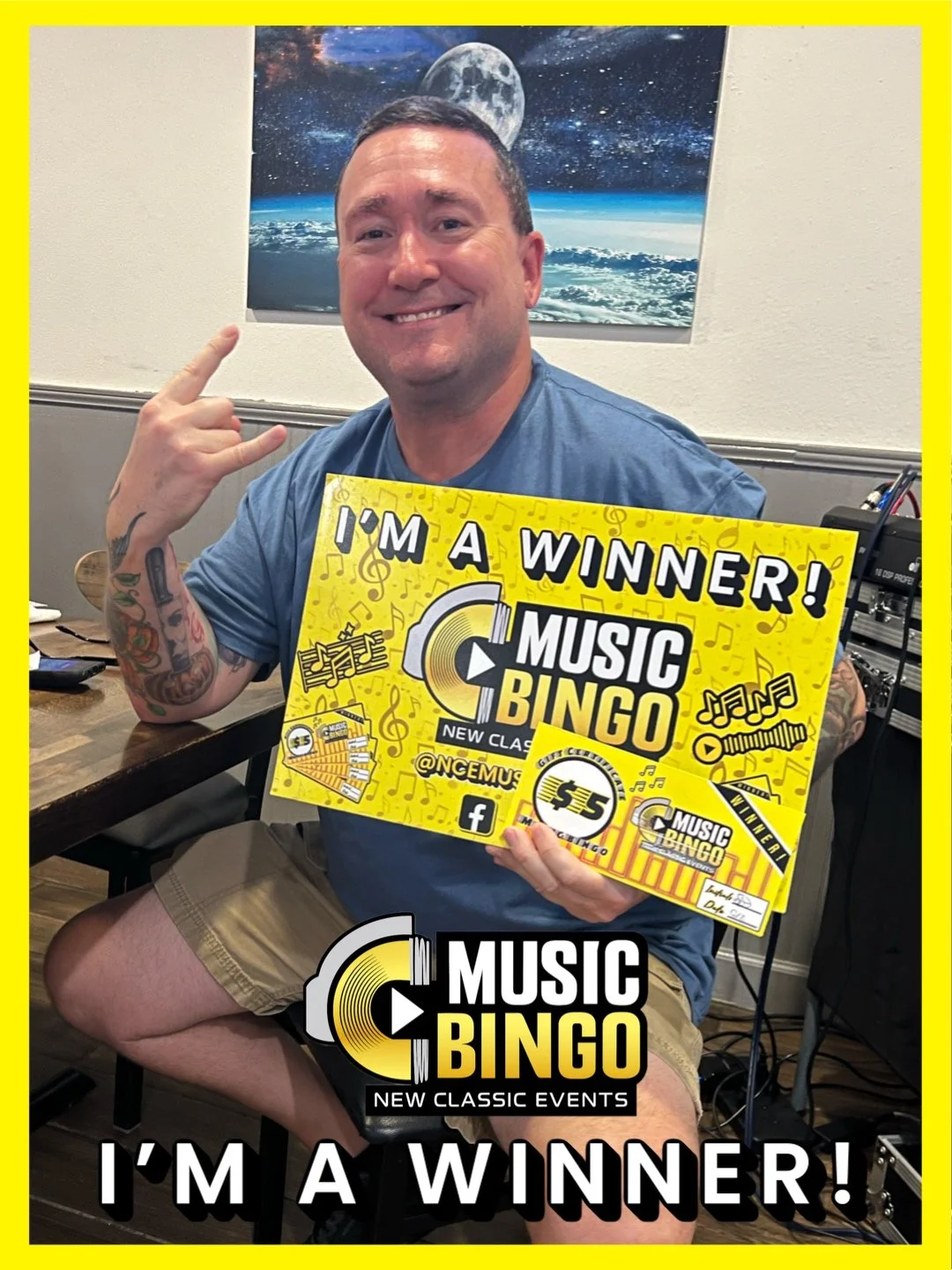 A man sitting indoors holding a yellow sign that reads 'I'm a winner! Music Bingo.' He is smiling and making a rock gesture with his right hand. There is a large poster of space and the moon on the wall behind him.