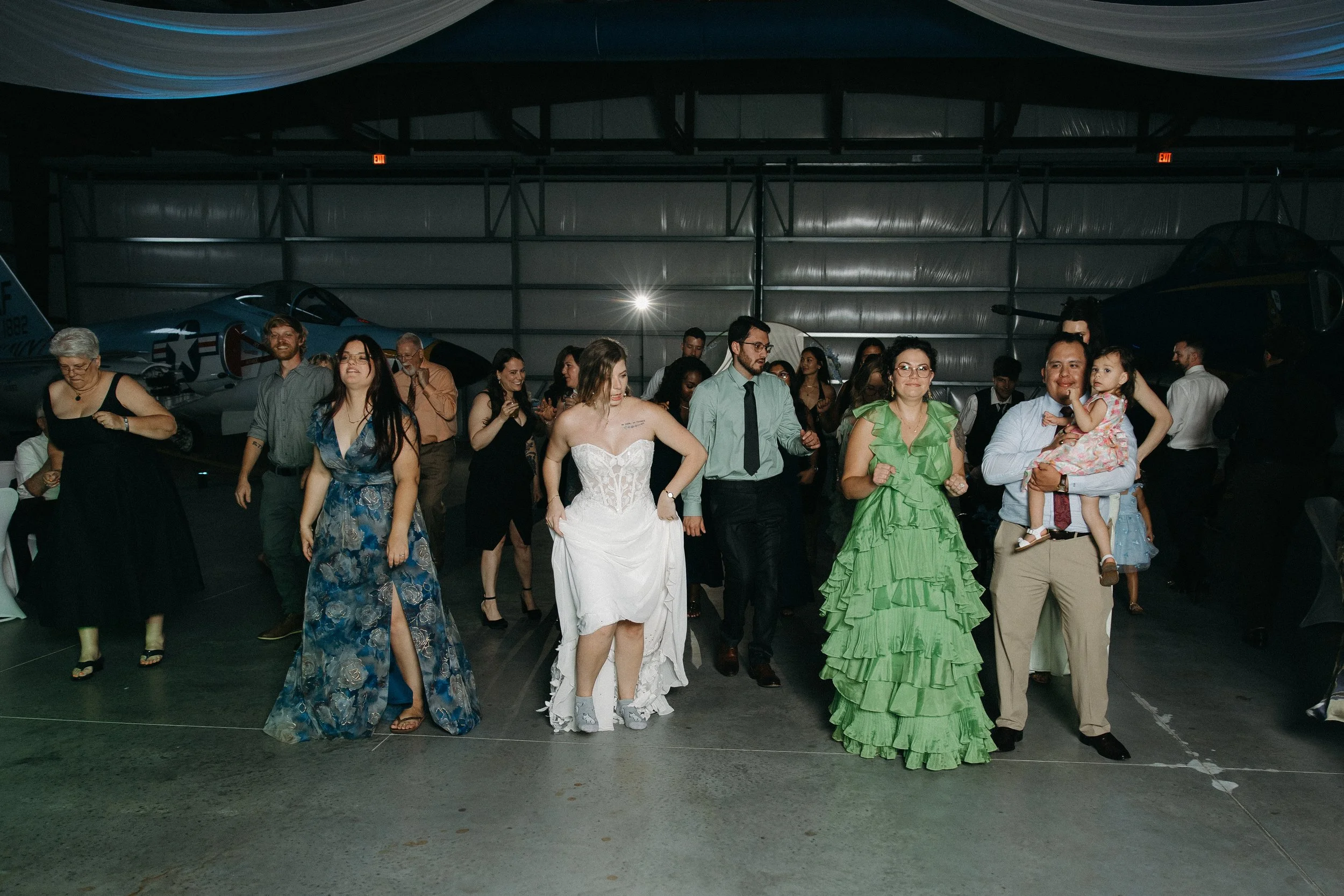 A diverse group of people dressed in formal attire dancing and celebrating in an indoor hangar with vintage aircraft in the background.