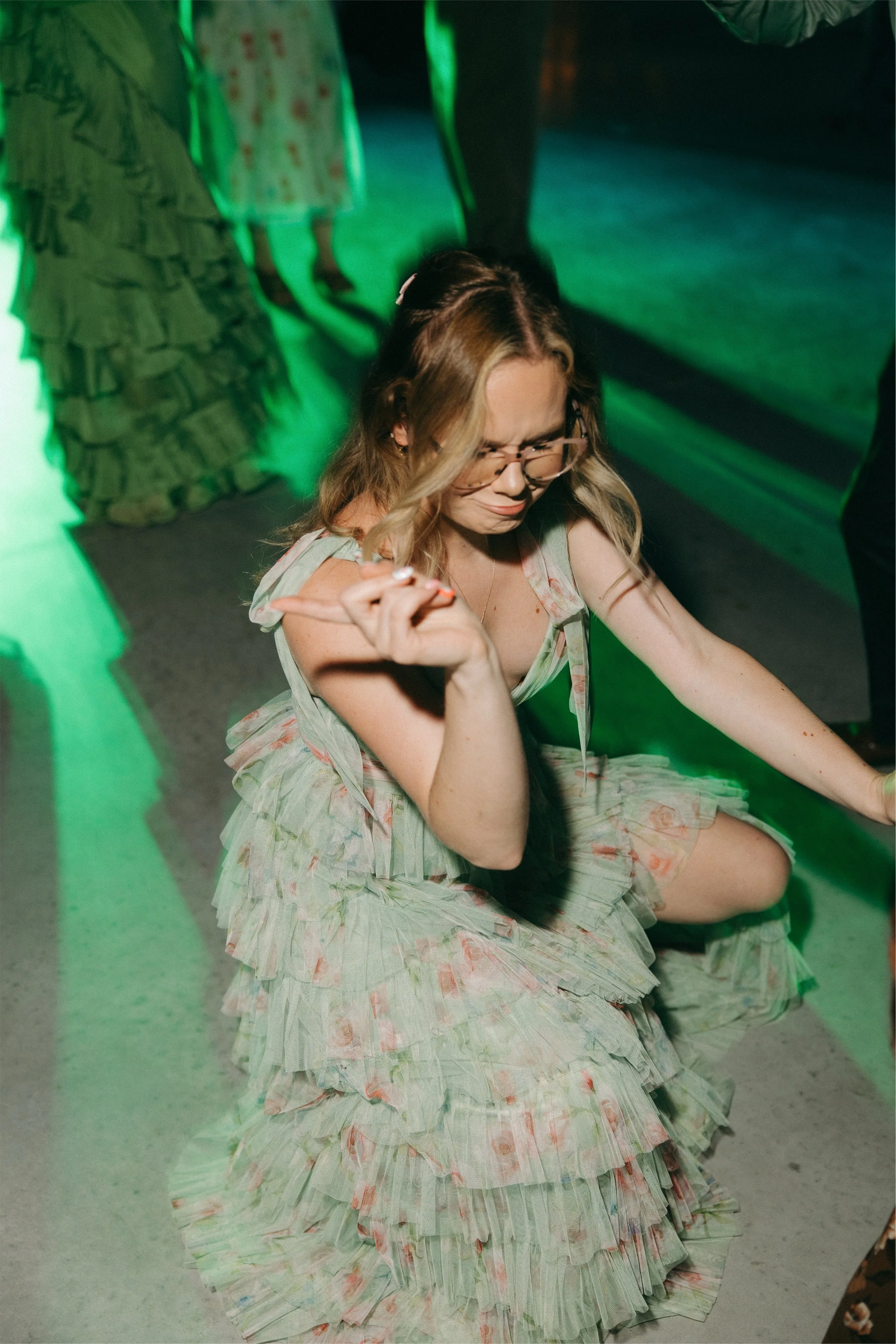 A woman with blonde hair wearing glasses and a floral layered dress is dancing in a dimly lit room with green lighting.