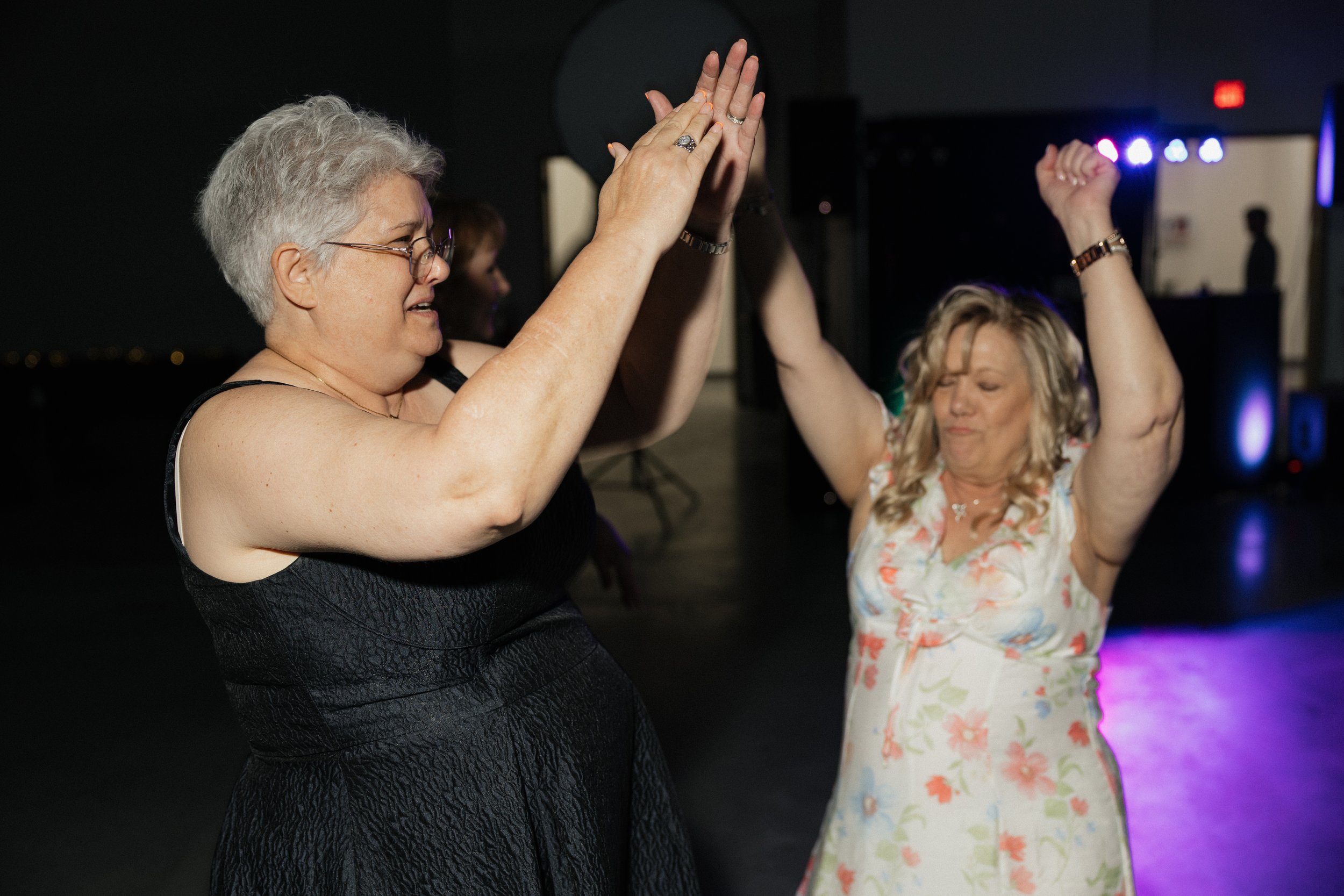 Two women dance with their hands raised, smiling, at a nighttime indoor event with colorful stage lighting.