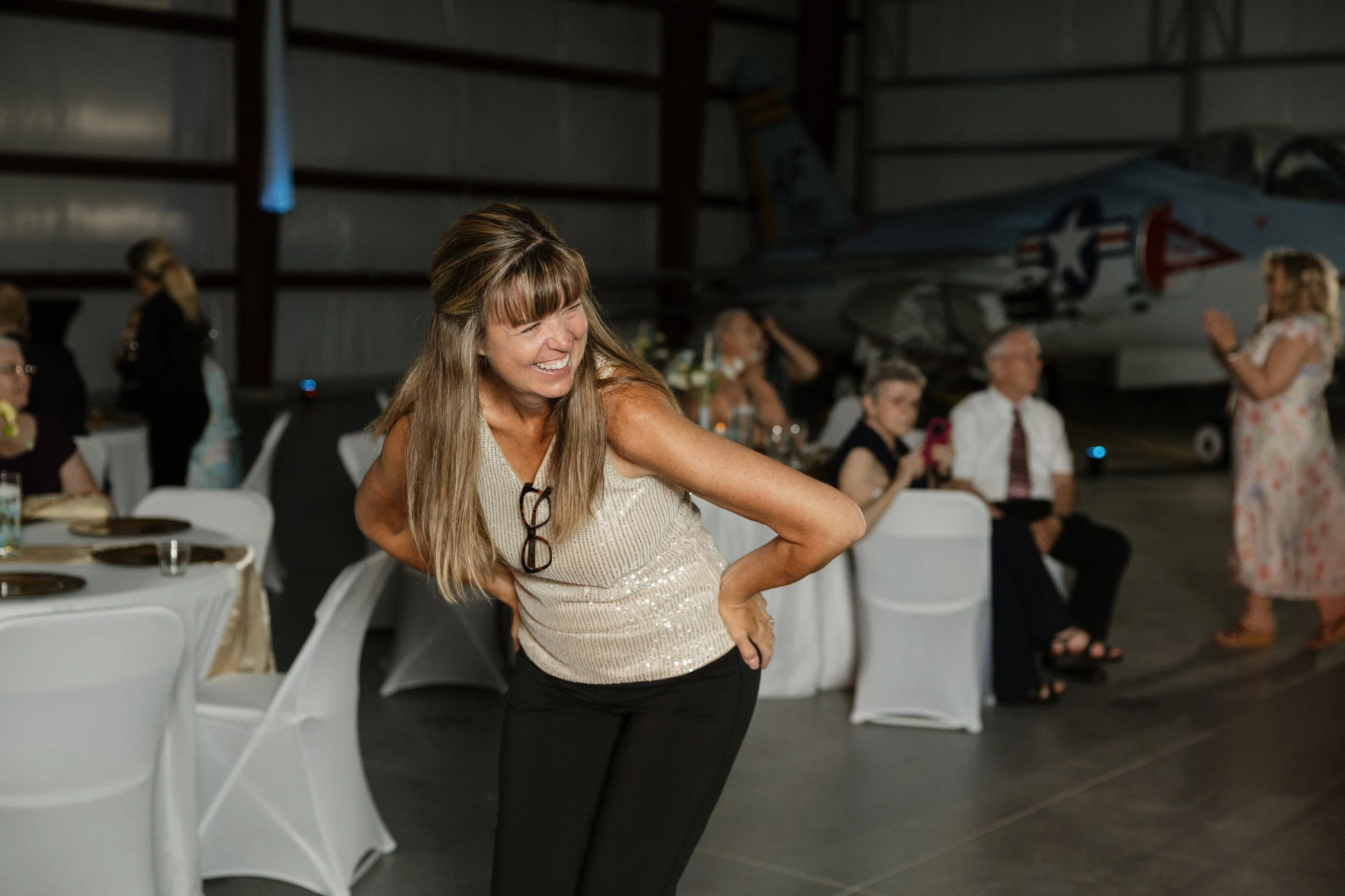 A woman with long hair smiling and leaning forward at a social gathering inside a hangar with a fighter jet in the background.