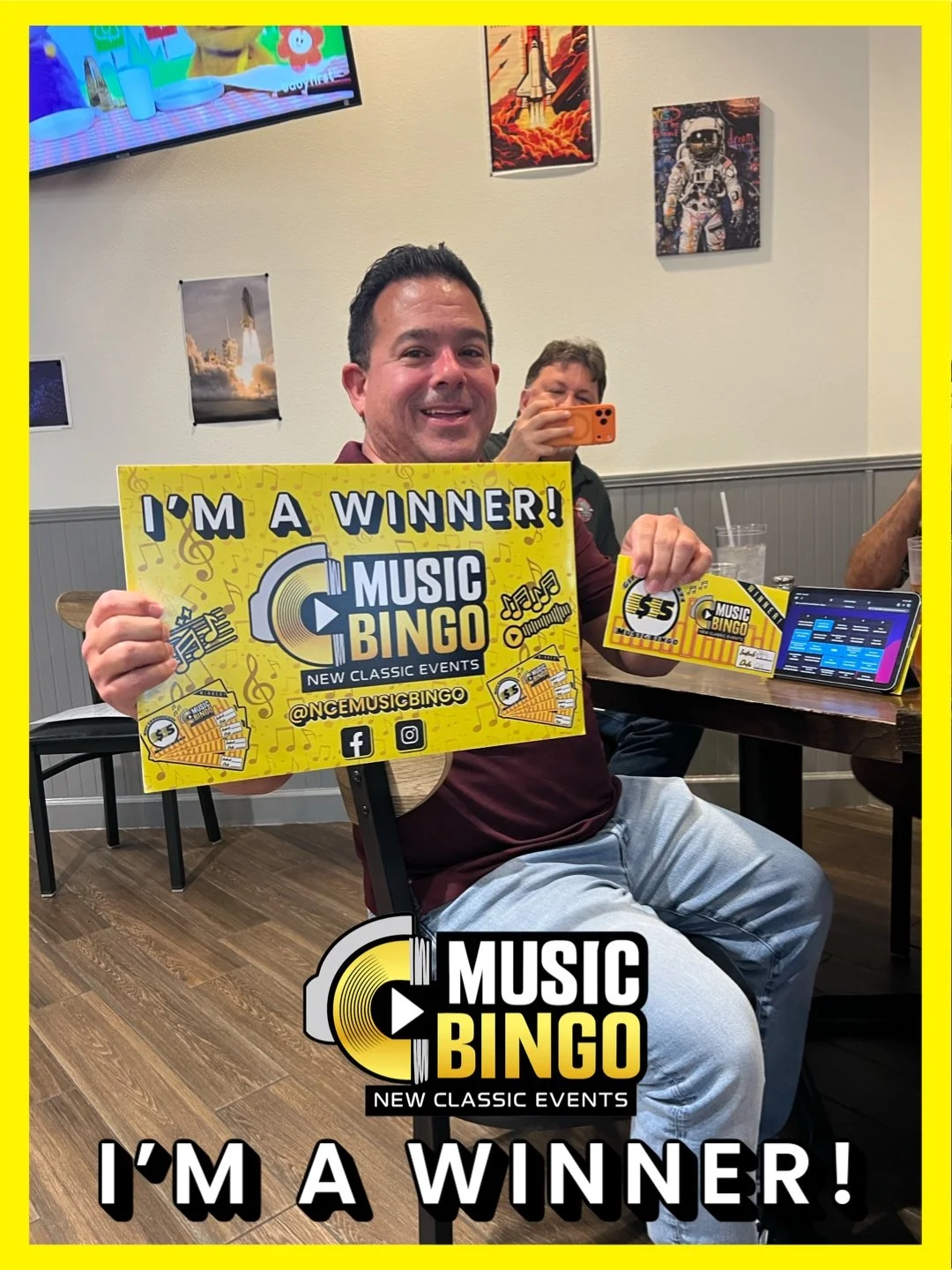 A man sitting at a table holding a large yellow sign that says "I'm a Winner! Music Bingo New Classic Events," with smaller flyers and a smartphone on the table. Another person in the background is taking a photo with a smartphone. The setting appear